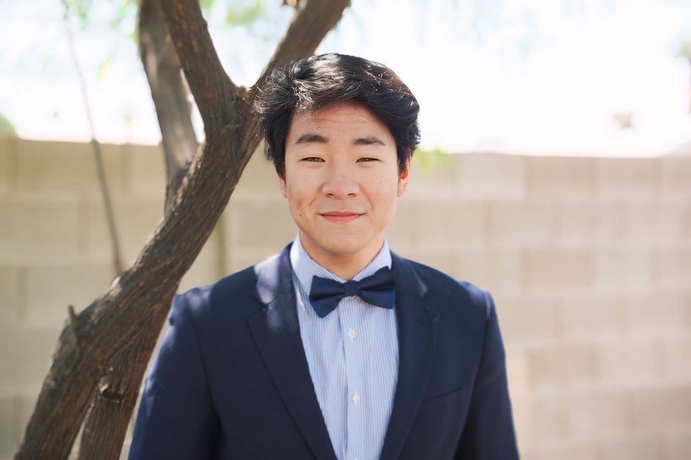 Young man dressed in a navy blue suit with a bow tie, standing outdoors near a tree and smiling.