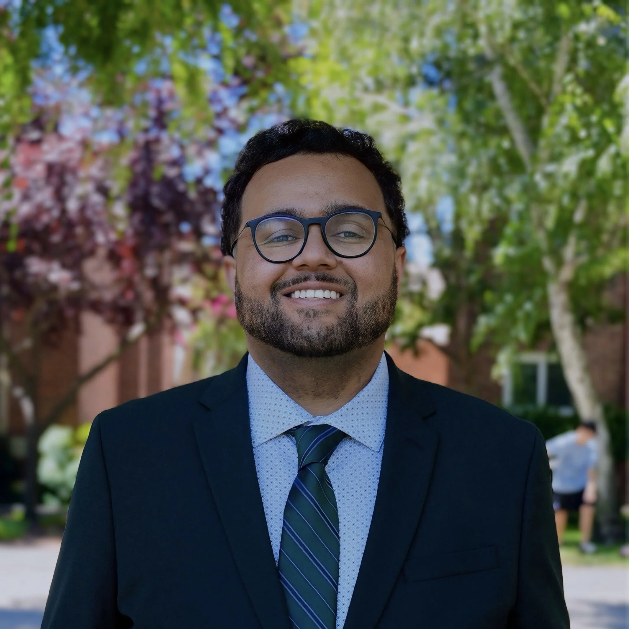 Man wearing glasses, a tan coat, and a hoodie, standing outdoors near water with a city skyline in the background.