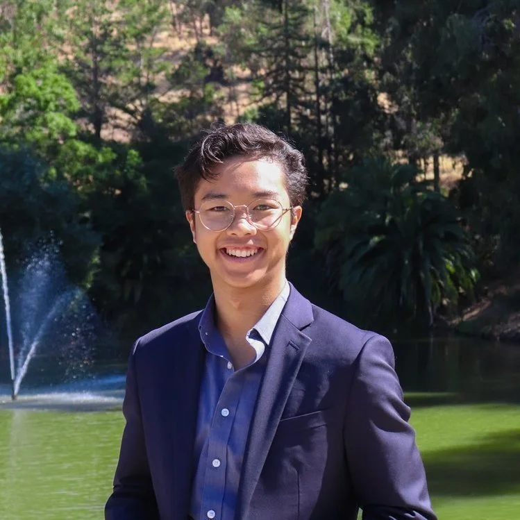 A young man in a navy suit and glasses smiling outdoors with a lake, fountain, and trees in the background.