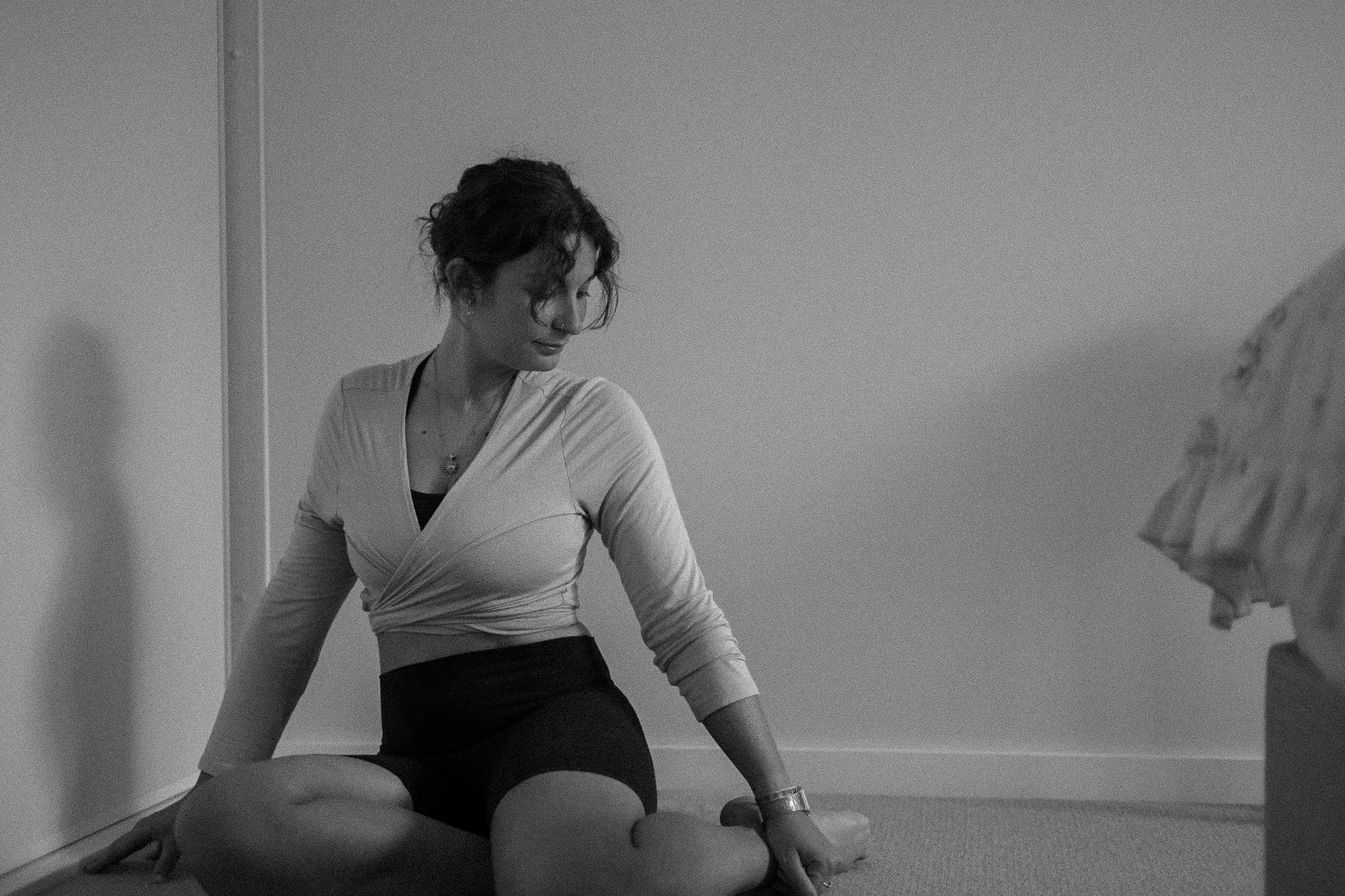 A woman sitting on the floor in a room, looking down, with her legs crossed and one hand resting on the floor. The room has plain walls and minimal decor.