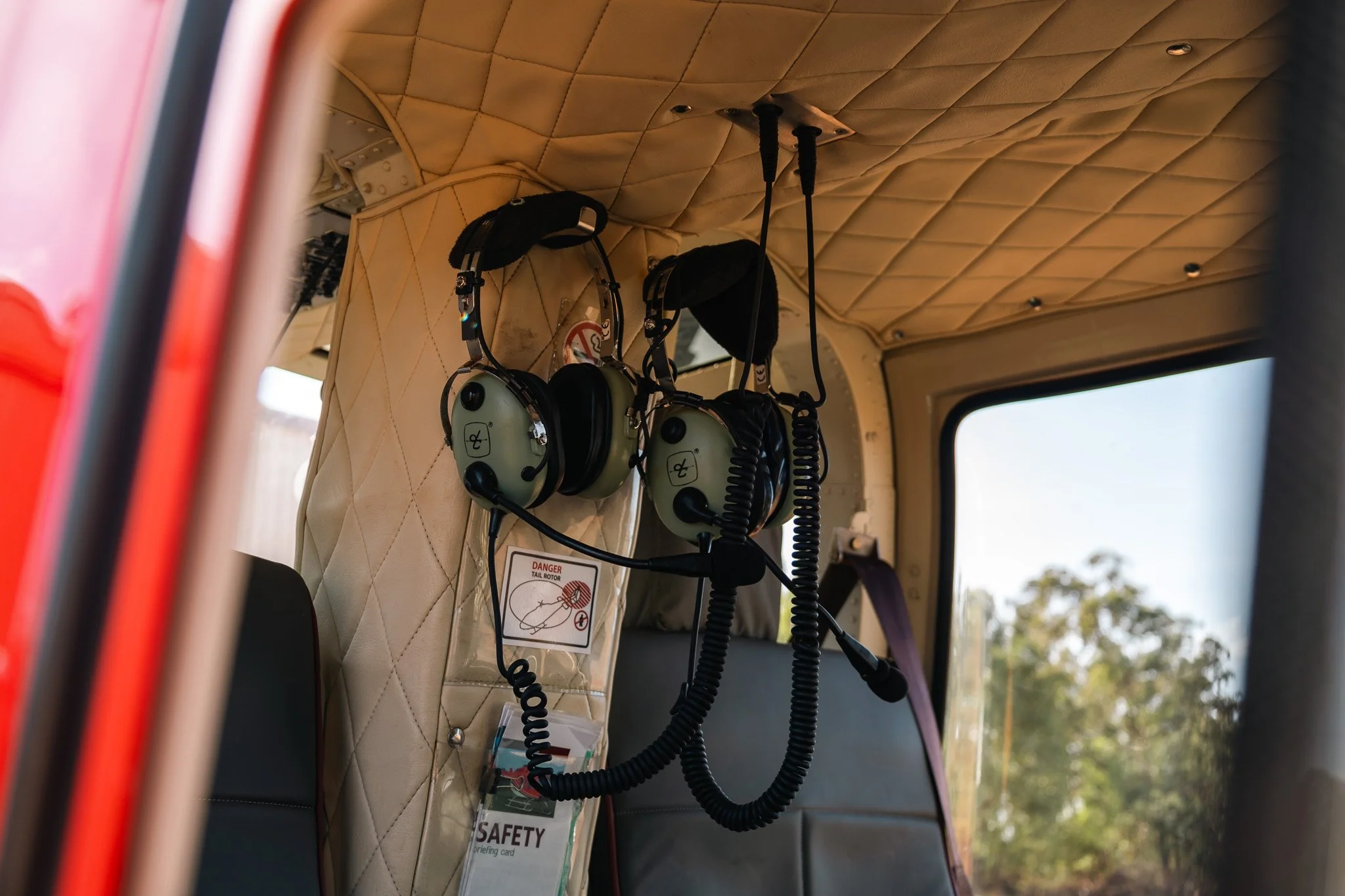 Two aviation headphones hanging from hooks inside a helicopter cockpit with a window showing trees outside.