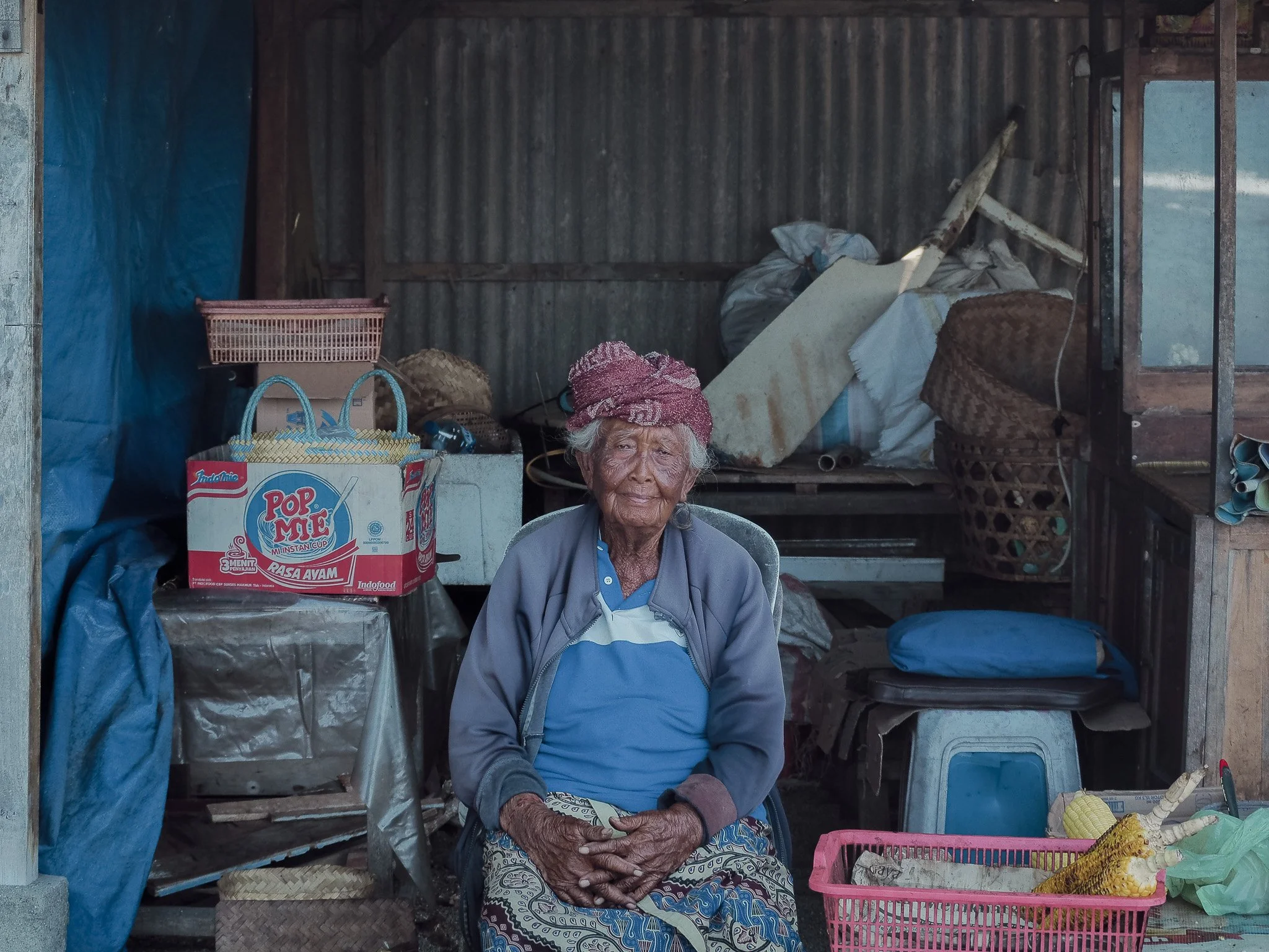 An elderly woman with a purple headscarf sitting in a small, rustic shop or stall with various household items and food products behind her.