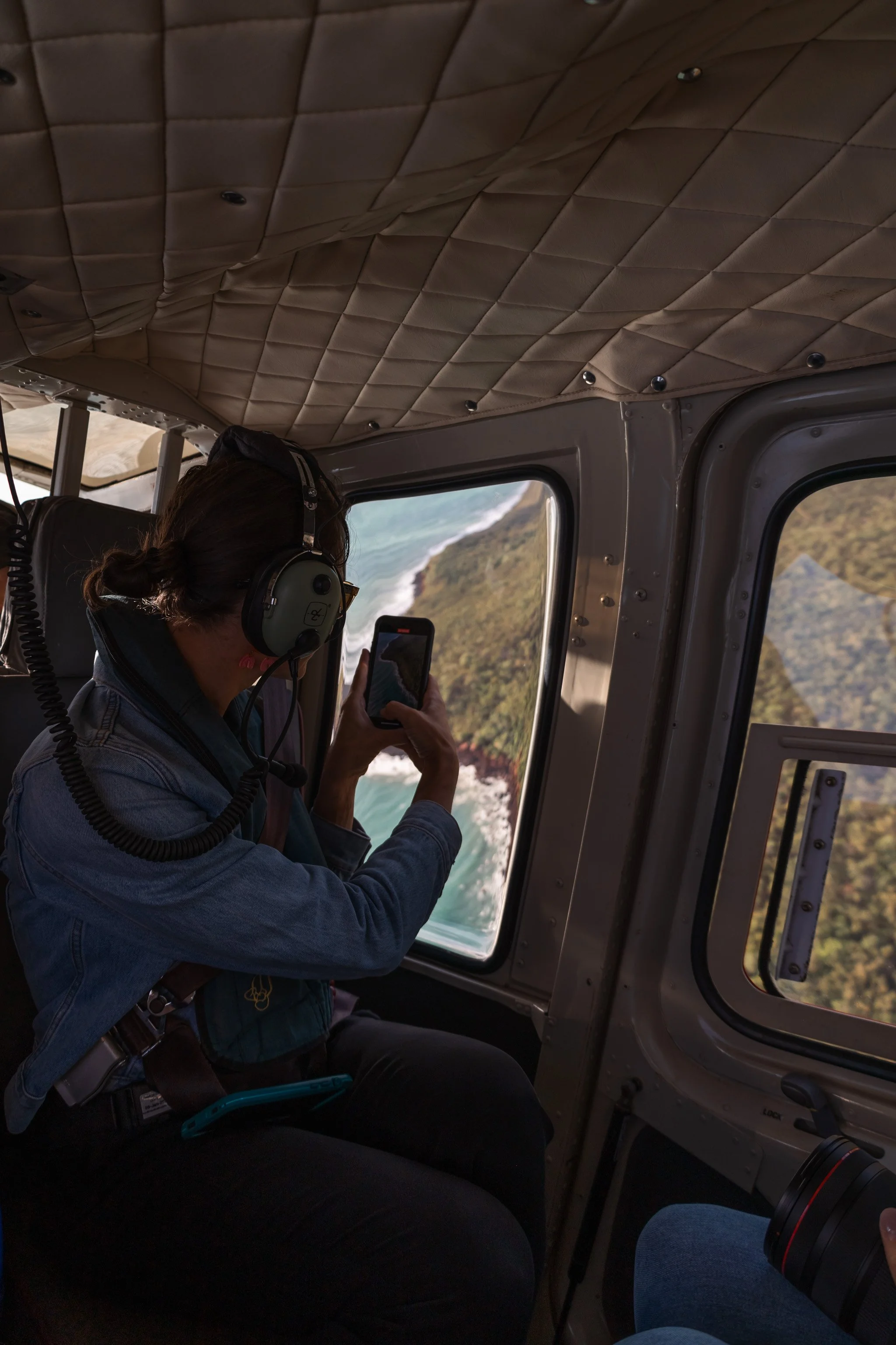A woman sitting inside a helicopter, taking a photo of the aerial view with her smartphone, with headphones on and wearing a denim jacket.