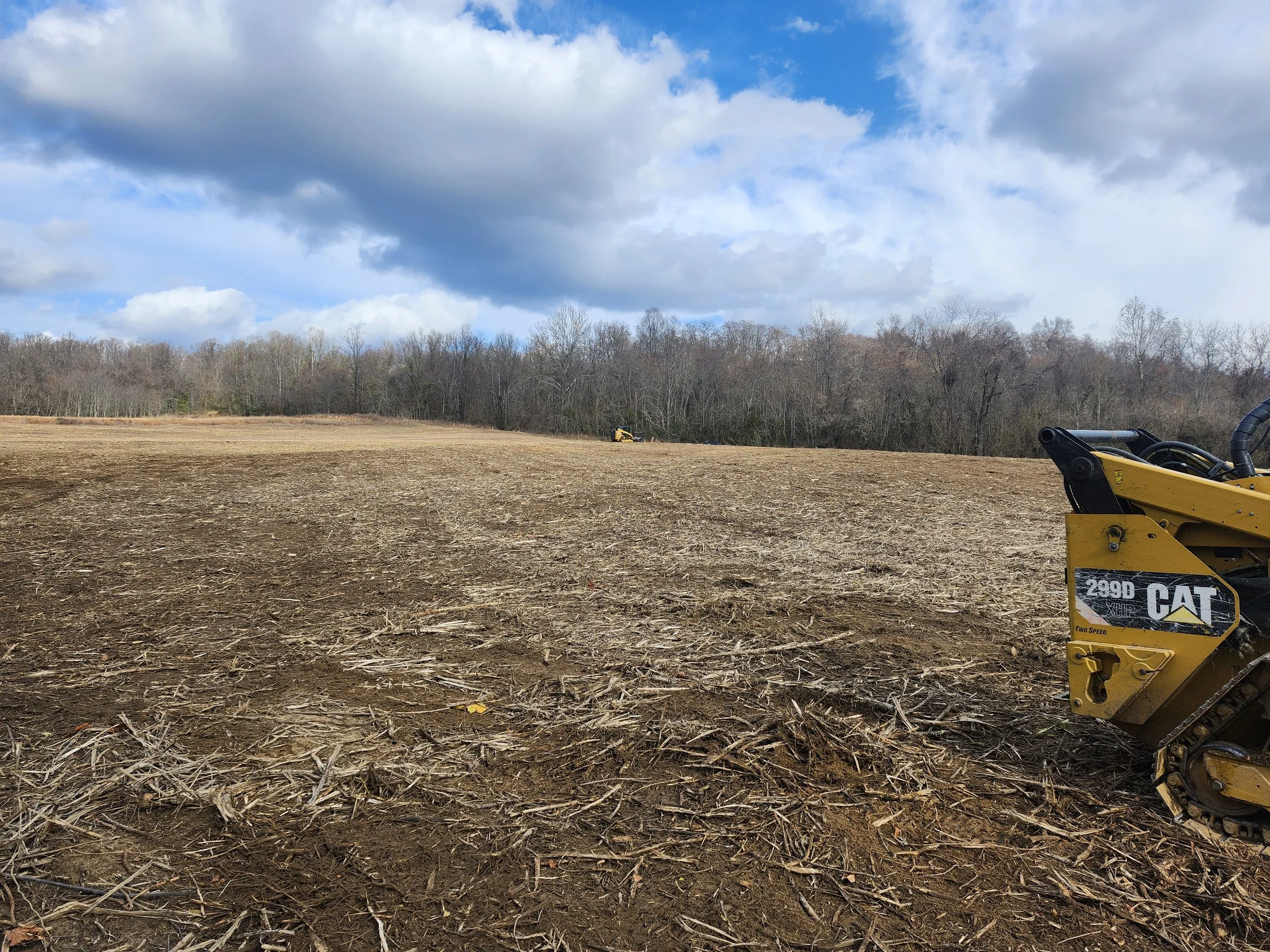 A large, open, freshly plowed field with a yellow CAT excavator on the right and another piece of construction equipment in the background near a tree line under a partly cloudy sky.
