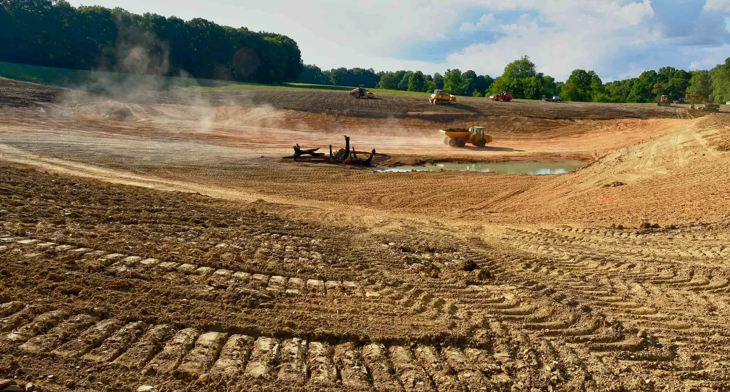 Excavated pond basin with heavy equipment and spoil piles under a blue sky.