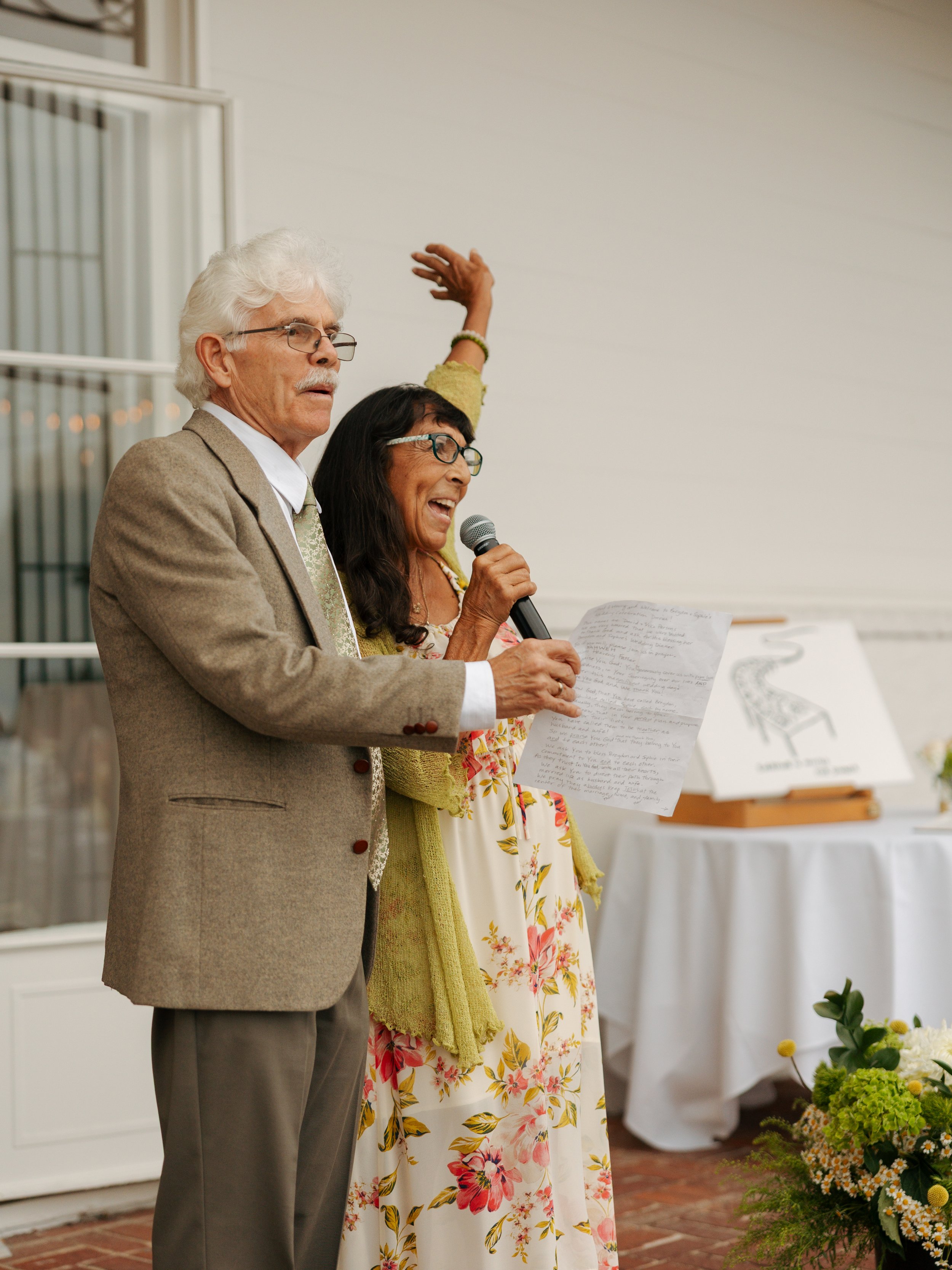 A couple giving a speech at an outdoor wedding reception, standing on a brick patio with floral decorations and potted plants nearby.