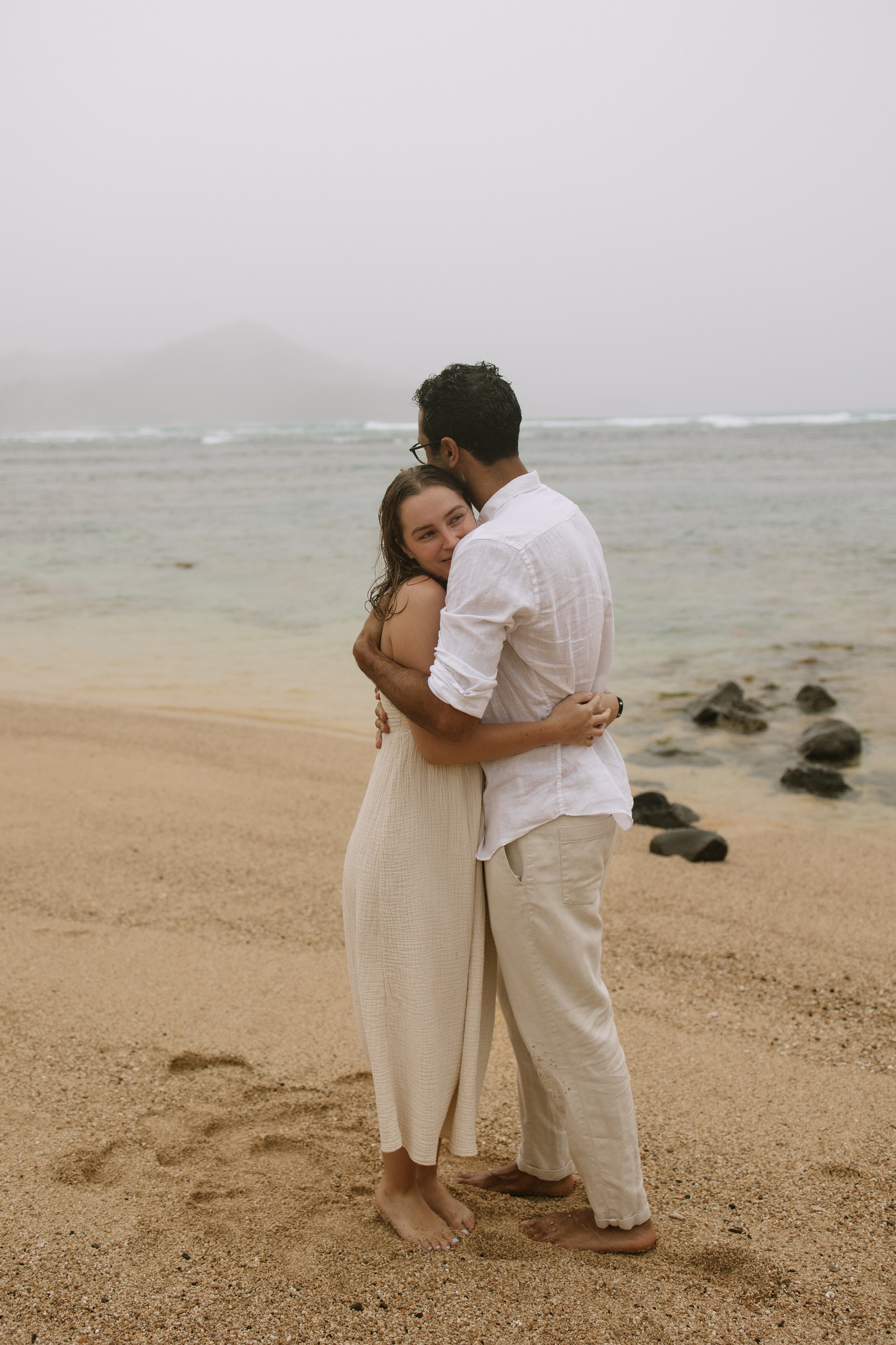 A couple hugging on a beach, with ocean and mountains in the background, overcast sky.
