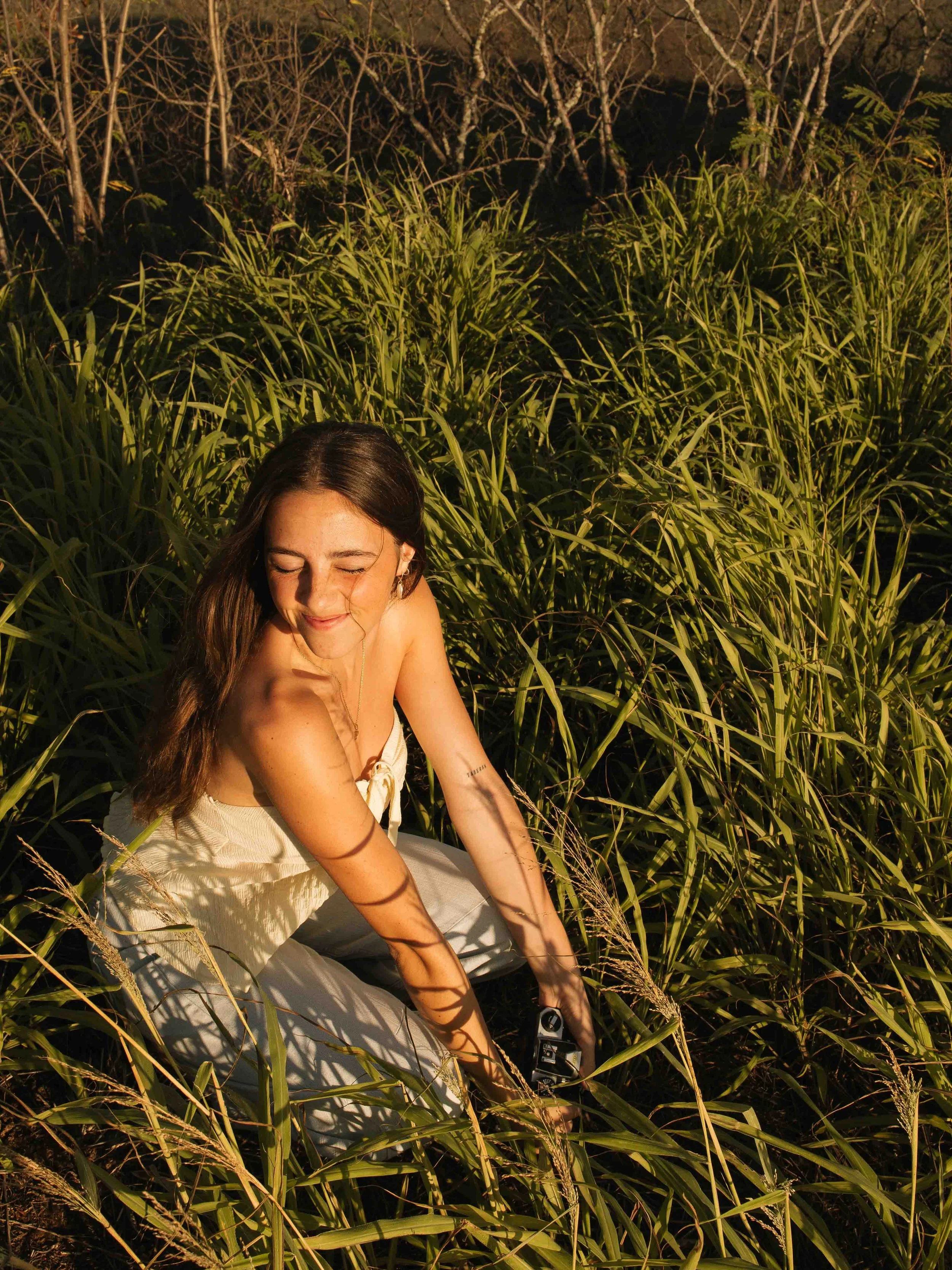 A woman with long brown hair smiling and squatting in a field of tall green grass, holding a camera, with sunlight casting shadows of the grass on her face.