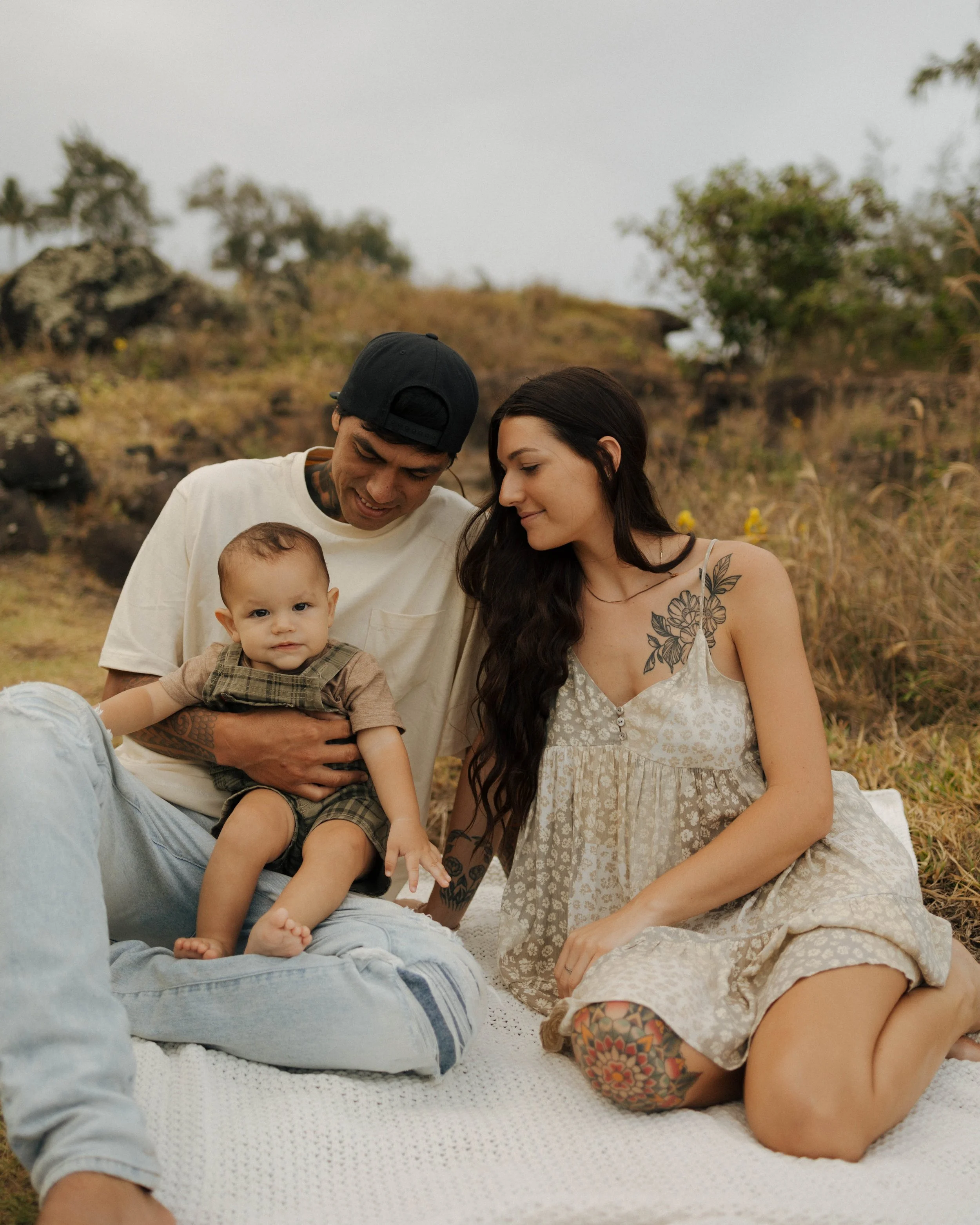 Family with a father, mother, and young child sitting on a blanket outdoors in a natural setting during daytime.