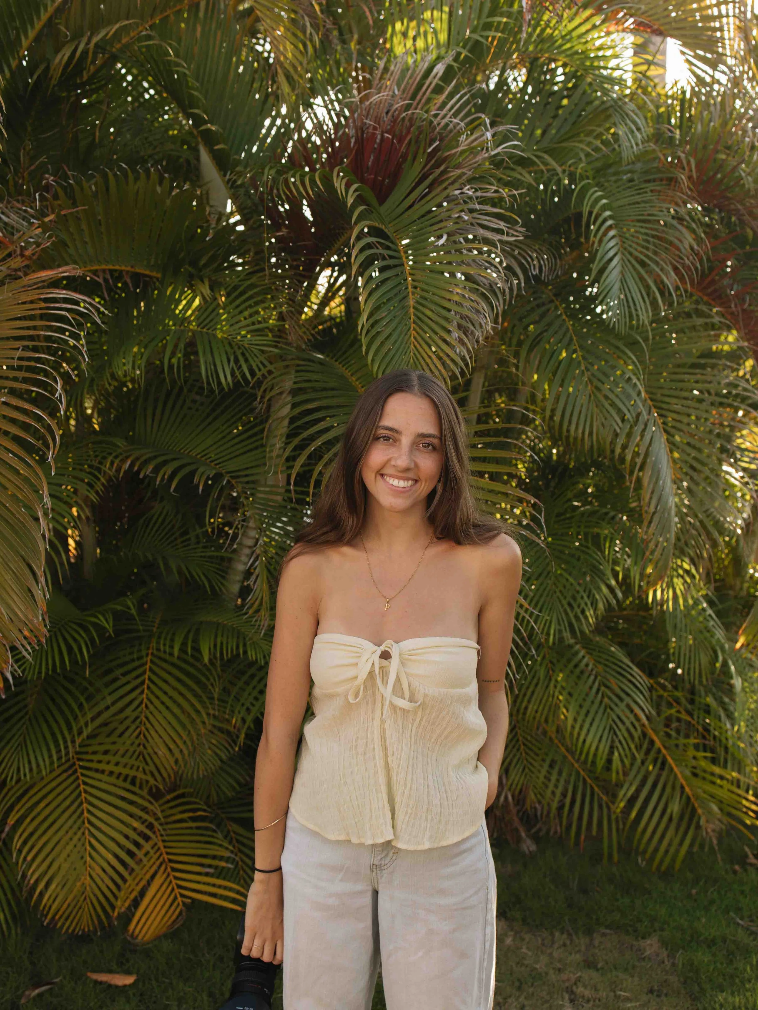 A young woman with long brown hair smiling outdoors in front of green and red palm leaves, wearing a strapless cream-colored top and light-colored pants.