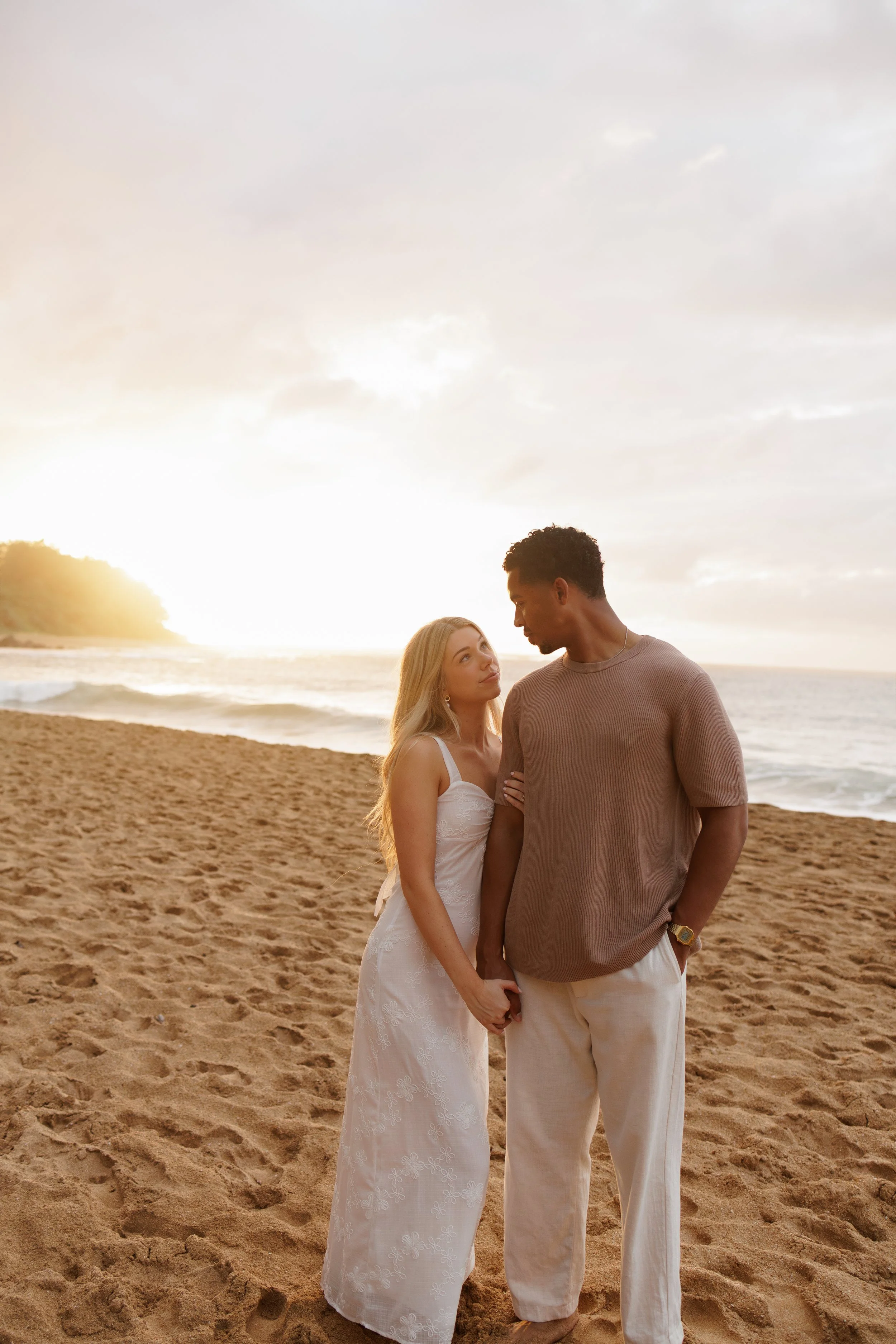 A couple hugging on a beach, with ocean and mountains in the background, overcast sky.