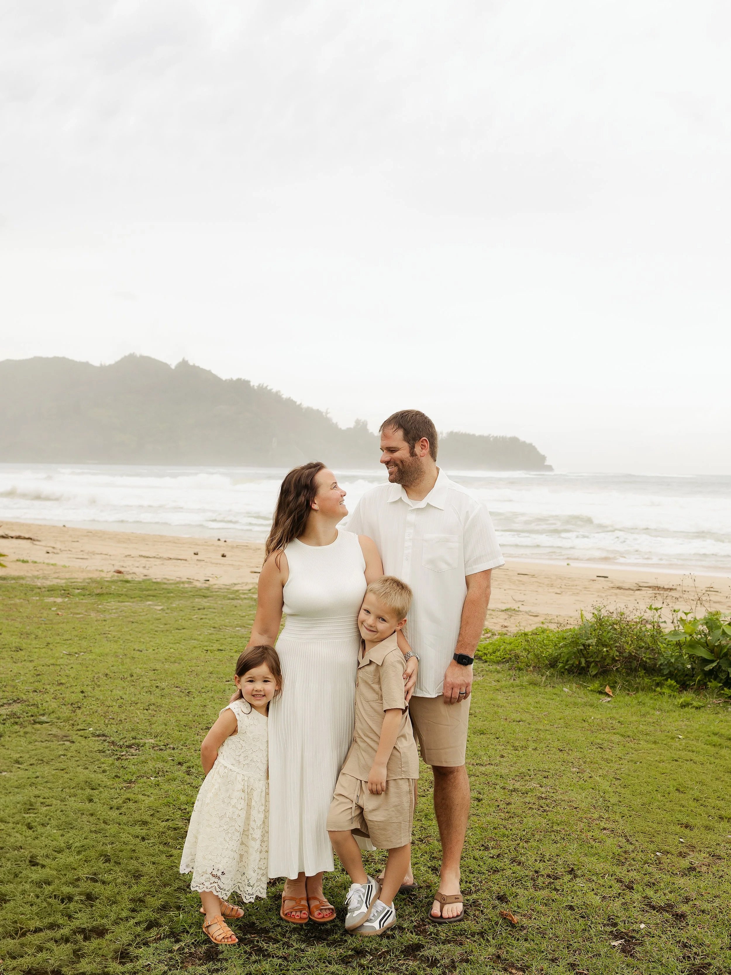 A family of five standing on a grassy area near the beach, with waves and a distant forested hill in the background. The family includes a woman in a white dress, a man in a white shirt and shorts, a girl in a lace dress, a boy in beige shorts and shirt, all smiling and looking at each other.