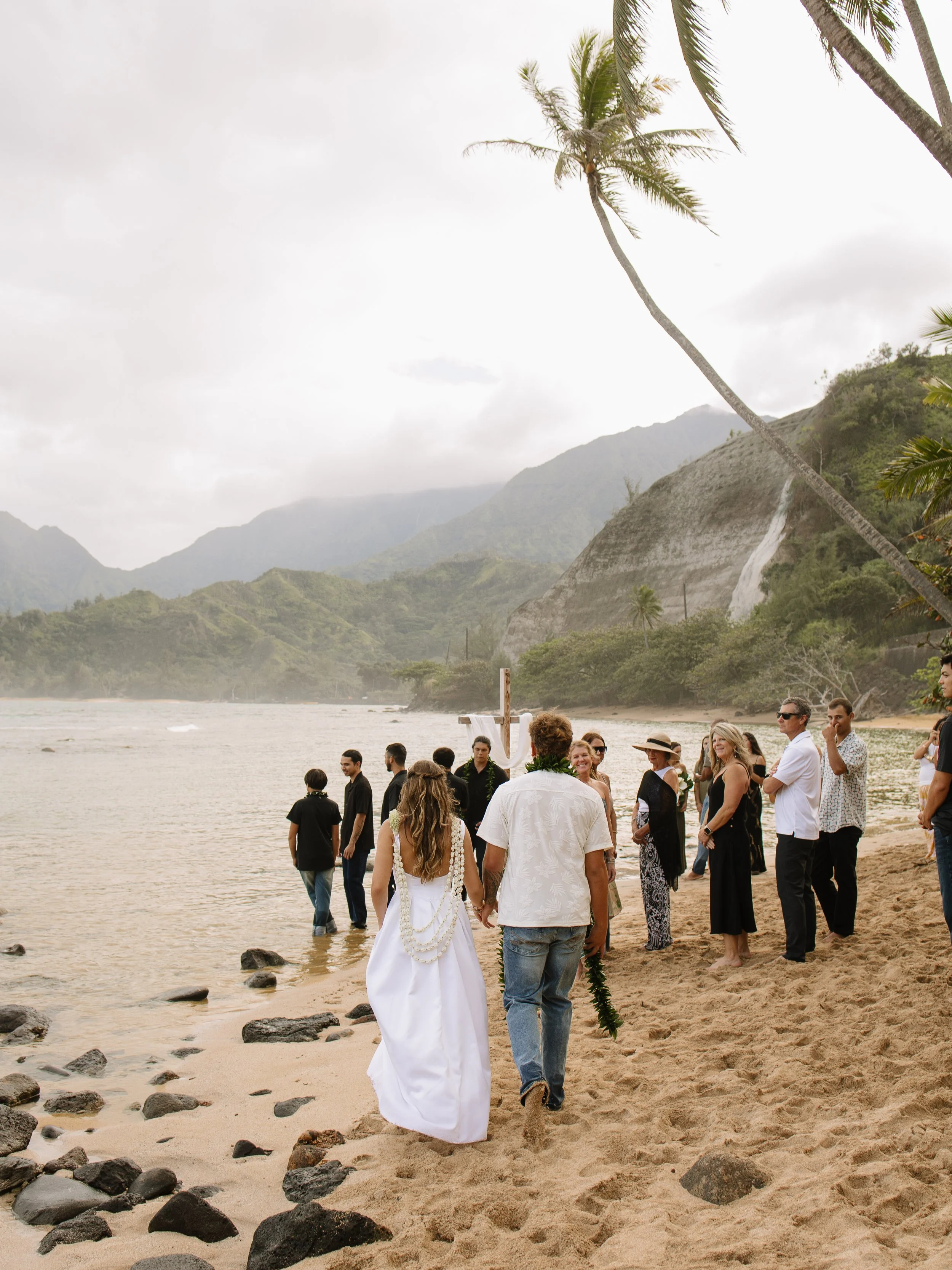 Couple walking hand-in-hand on a beach during a wedding ceremony, with a group of people witnessing by the water, surrounded by palm trees and lush green mountains under a cloudy sky.