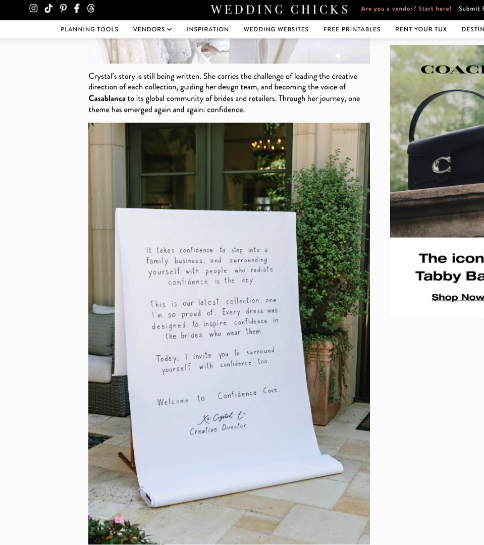 A white poster with handwritten text by JINA KOH Calligraphy is displayed , leaning against a stand. It is positioned on a paved surface with some greenery nearby, including potted plants and bushes.