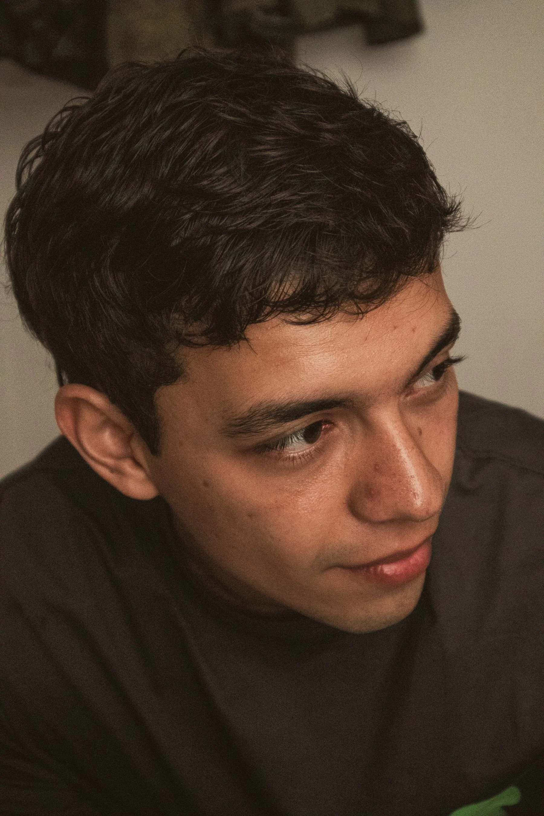 Close-up of a young man with short, wavy black hair, tan skin, and wearing a black shirt, looking down and smiling slightly.