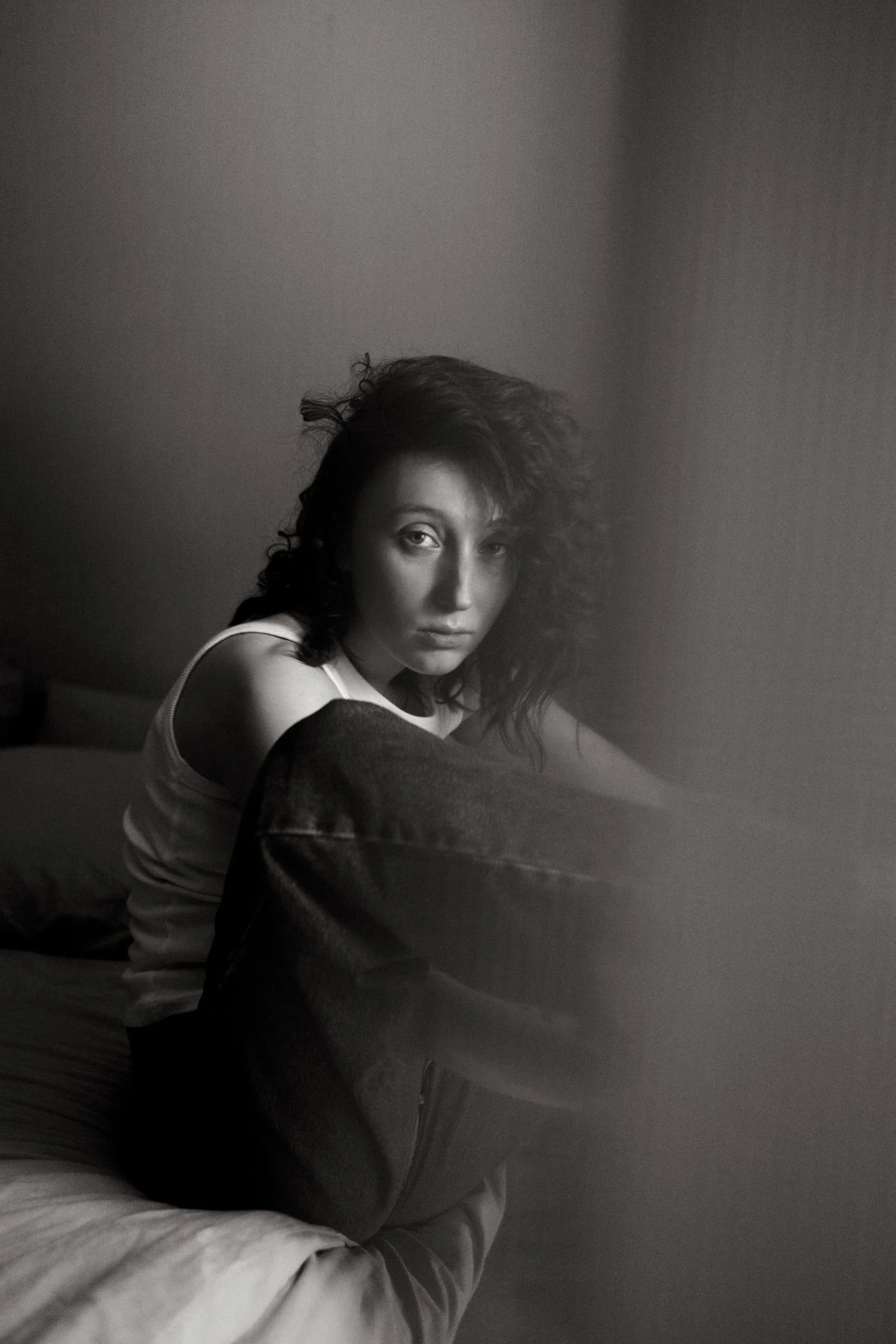 A black and white photo of a woman with curly hair, sitting and looking at the camera, with the bottom half of her body visible and some furniture around her.