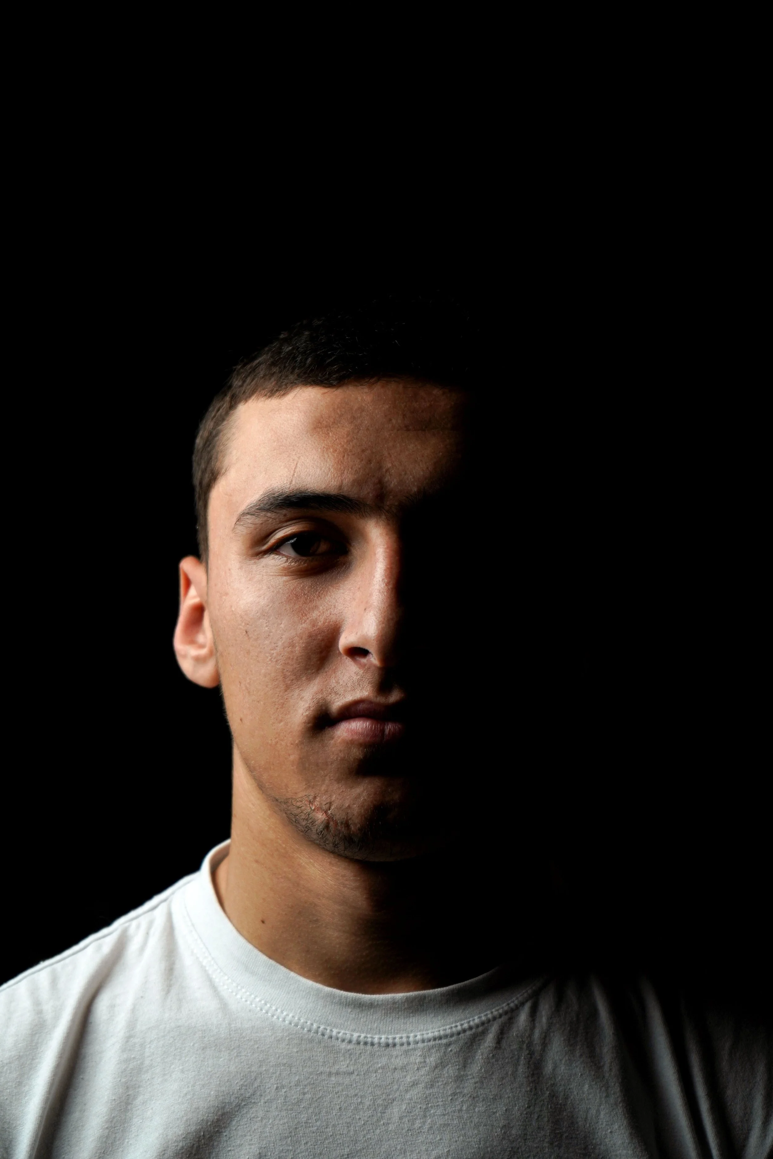Close-up of a young man's face with short dark hair, visible eye, and a slight beard, wearing a white shirt against a dark background.