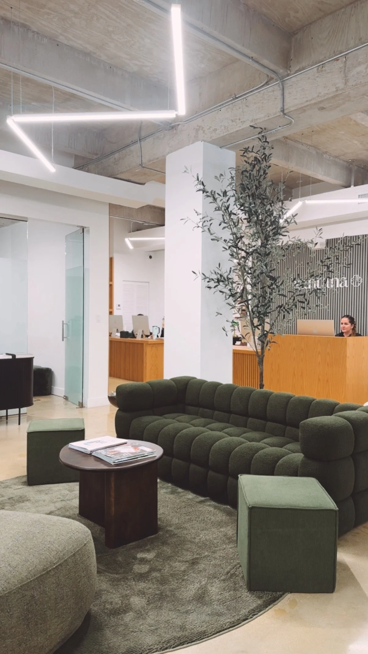 Modern lobby or reception area with green tufted sofa, a small round table with magazines, a green ottoman, and a grey armchair. There is a tall potted plant next to a white column and a wooden reception desk with a woman behind it. The ceiling is exposed concrete with modern LED lighting fixtures.