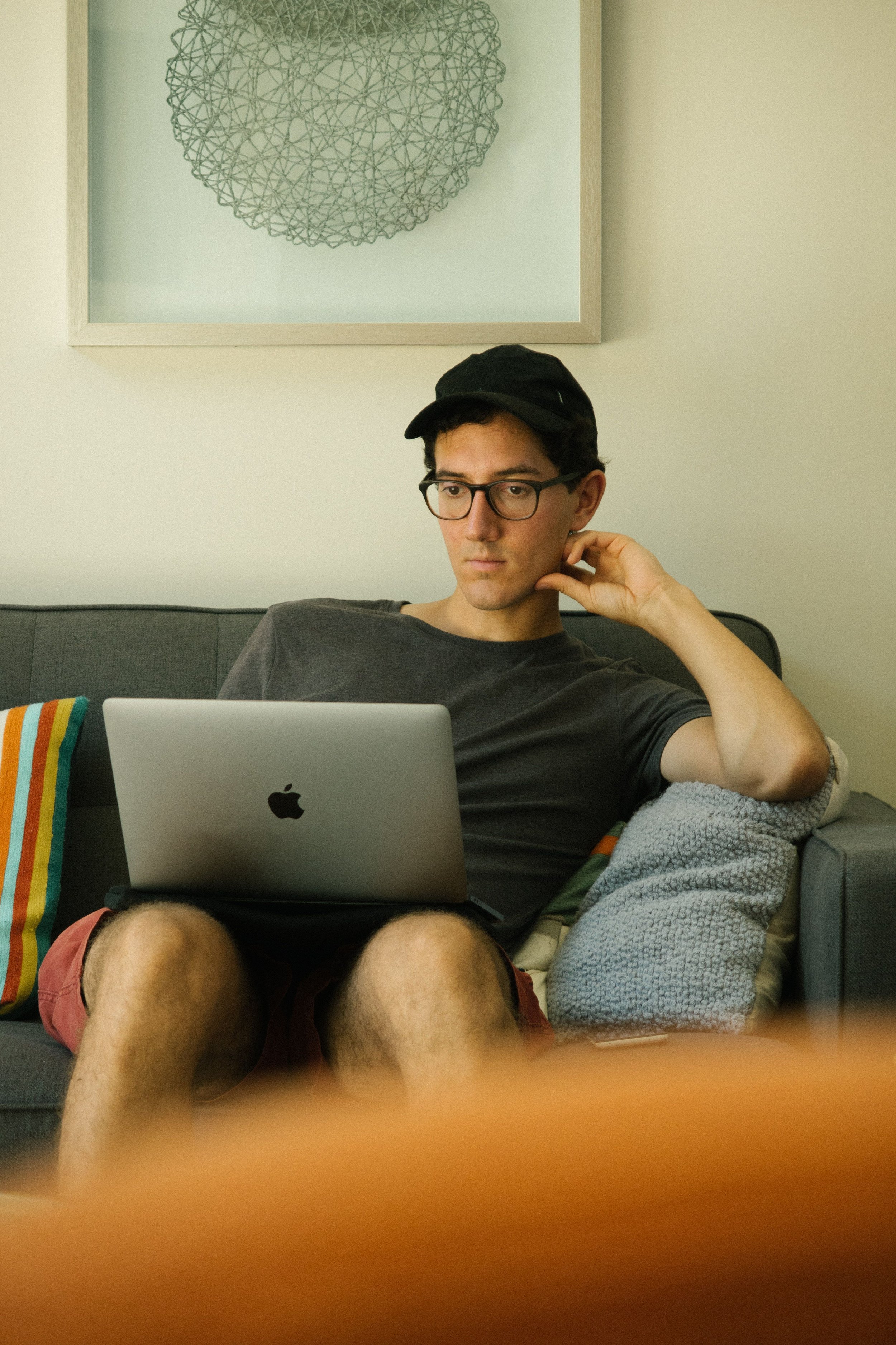A young man with glasses, wearing a black cap and a gray t-shirt, sitting on a couch with a laptop on his lap. His left hand is touching his neck, and he appears focused on the screen. Behind him is a light-colored wall with a framed abstract art piece, and a multicolored striped pillow is on the left side of the couch. A blanket is draped over his legs, and to his right, part of his body is visible.