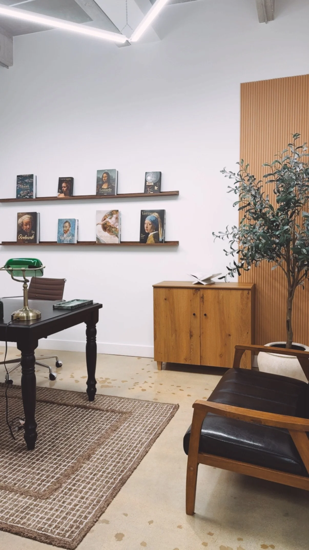 Interior of an office or study room with a black desk, green banker lamp, brown chair, wooden cabinet, potted plant, and a wall with books on two shelves featuring famous art and historical portraits.