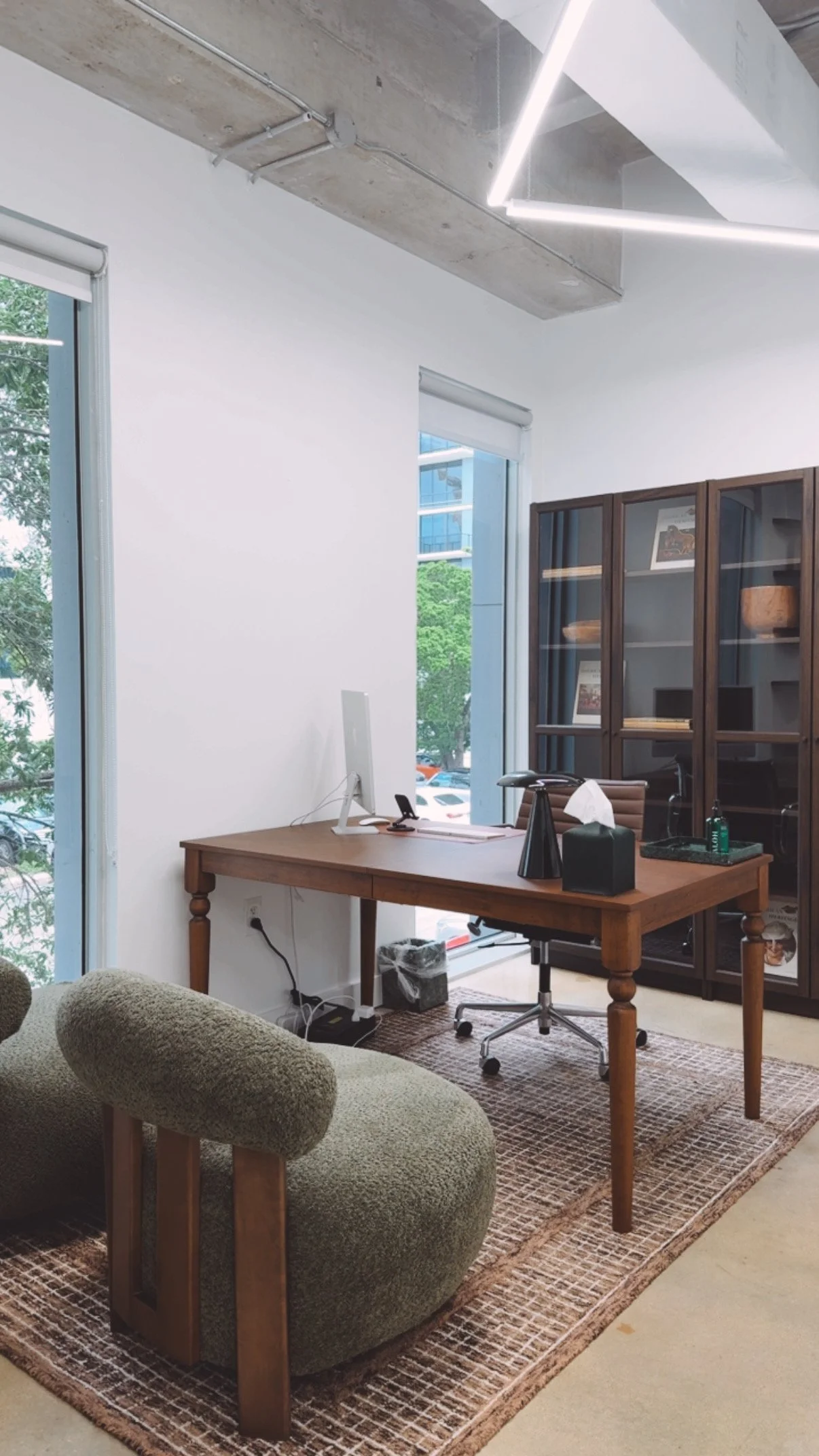 A modern office with a wooden desk, swivel chair, green upholstered armchair, large windows, a dark wood cabinet, and a concrete ceiling with LED lighting.