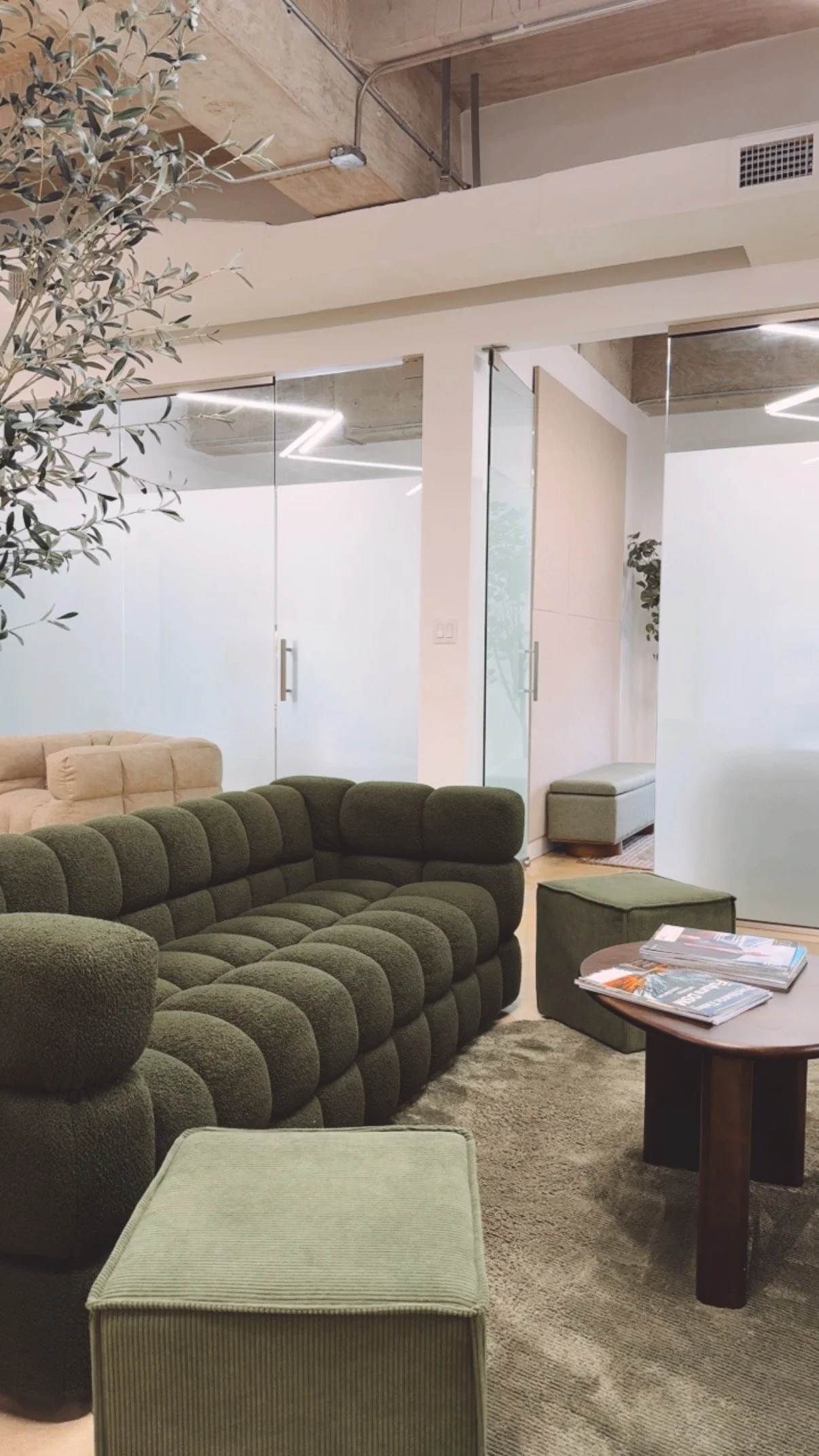 Modern lounge area with green tufted sofa, beige armchair, ottoman, and coffee table with magazines. Glass doors and minimalist decor, including a potted plant, are visible.