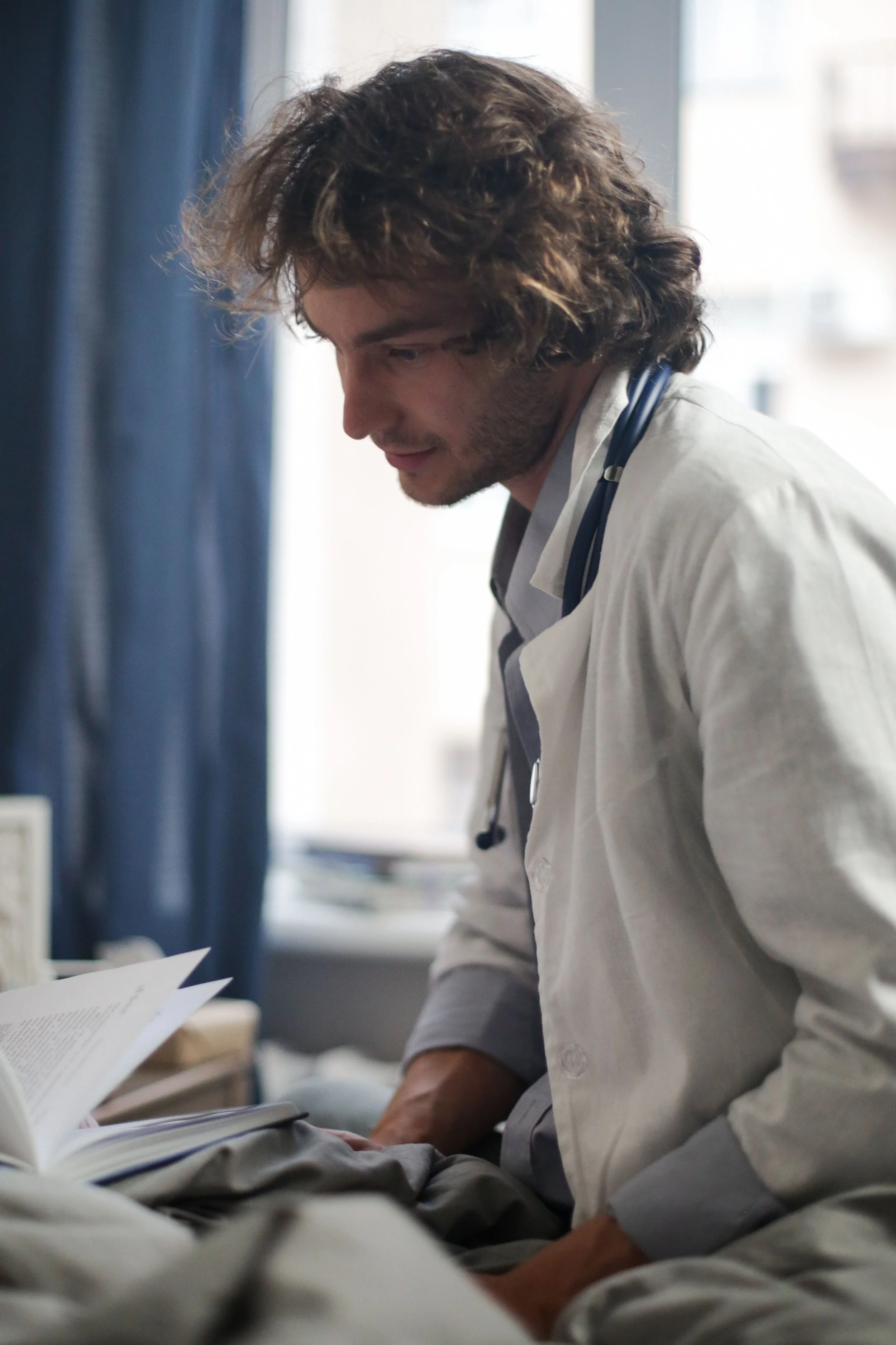 A man with curly brown hair and a stethoscope around his neck, wearing a white doctor coat, leaning over a patient, looking at papers on a desk in a clinical setting with natural light from a window.