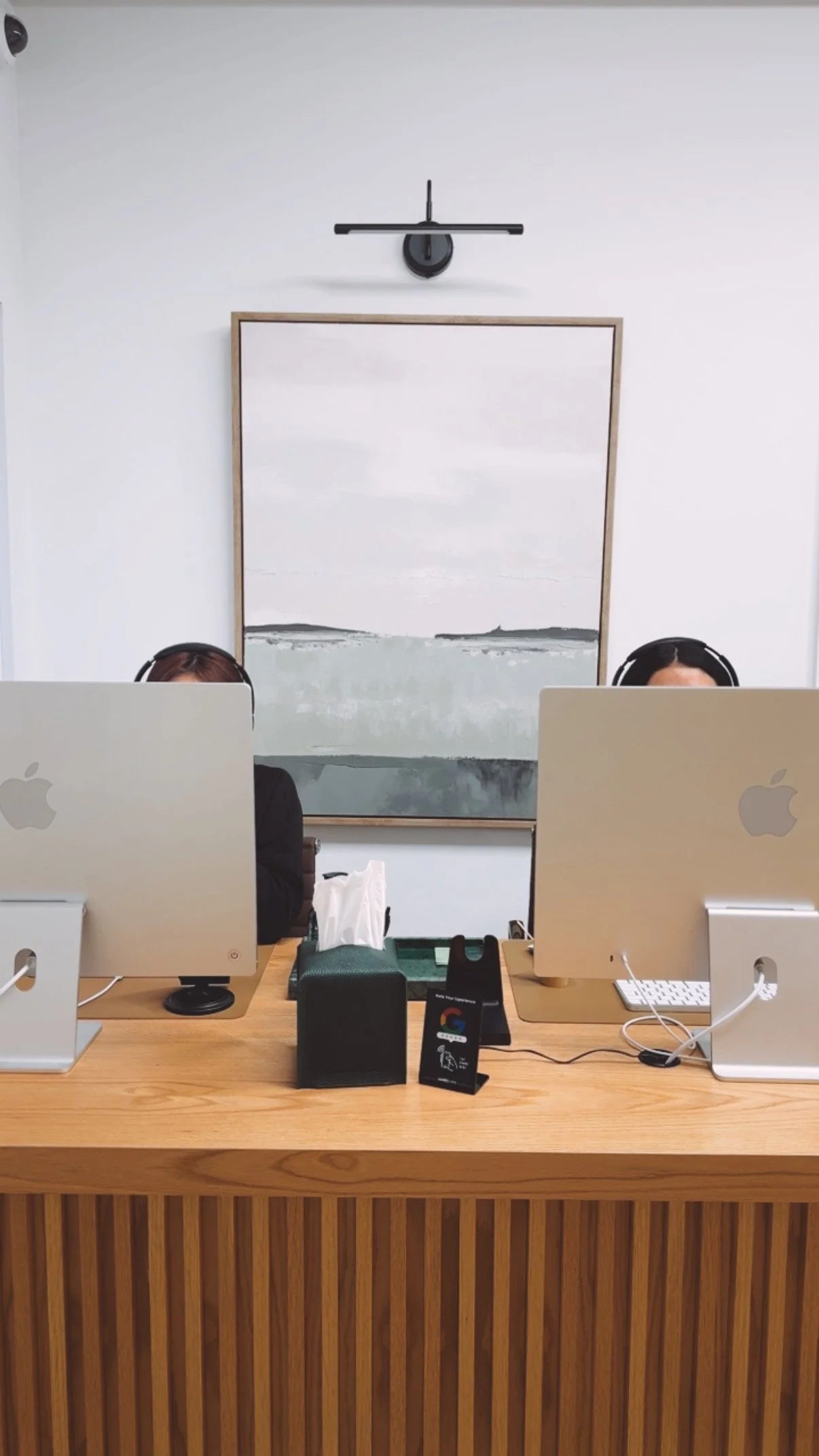An office workspace with a wooden front desk, two Mac computers, tissue box, tissue pack, and a Google card holder on the desk. Two people are partially visible working on computers, with a whiteboard and a black wall-mounted light fixture in the background.