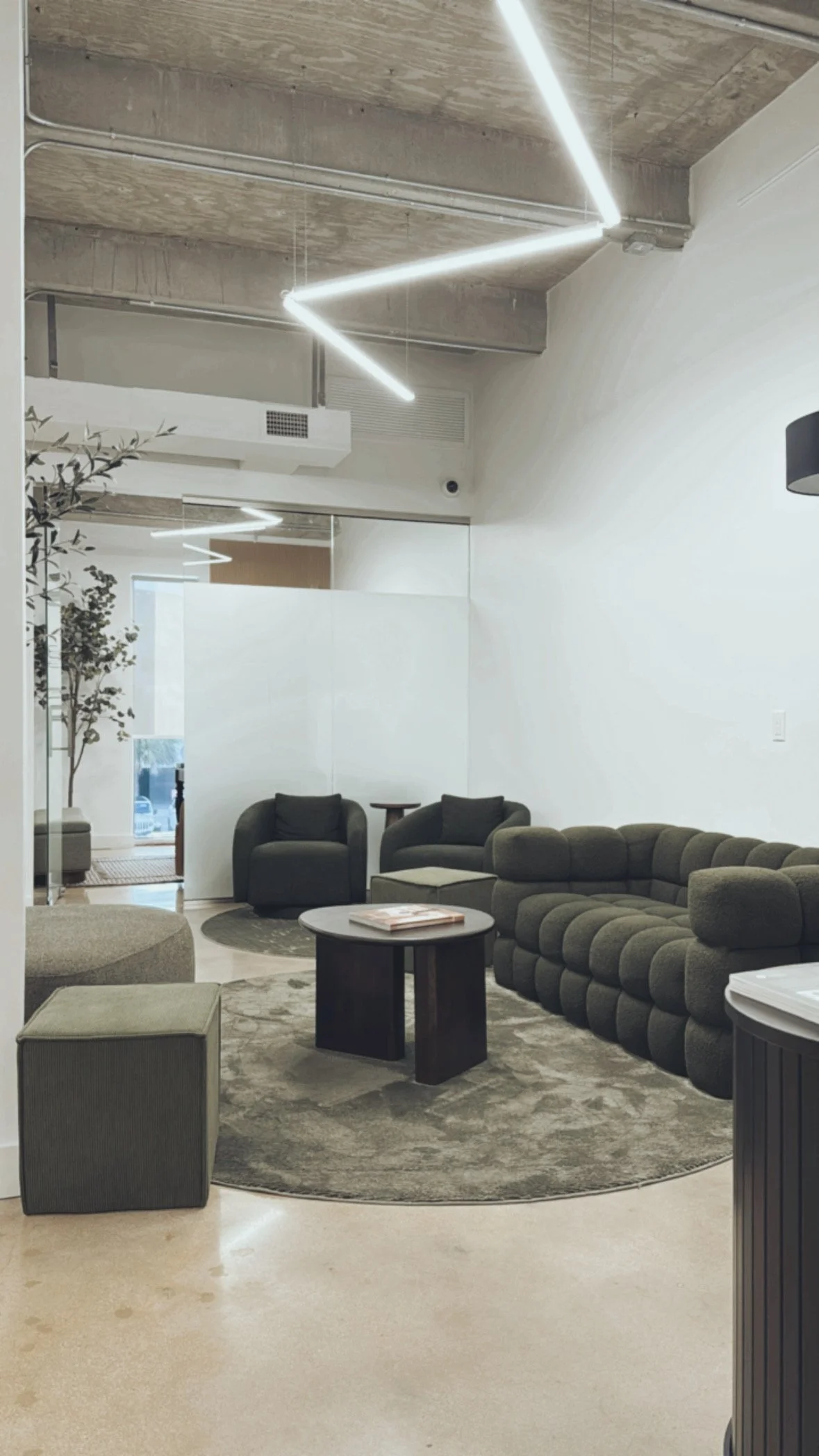 Modern lounge area with black and grey couches, a round black coffee table, a patterned grey rug, potted trees, and minimalist lighting fixtures on the ceiling.