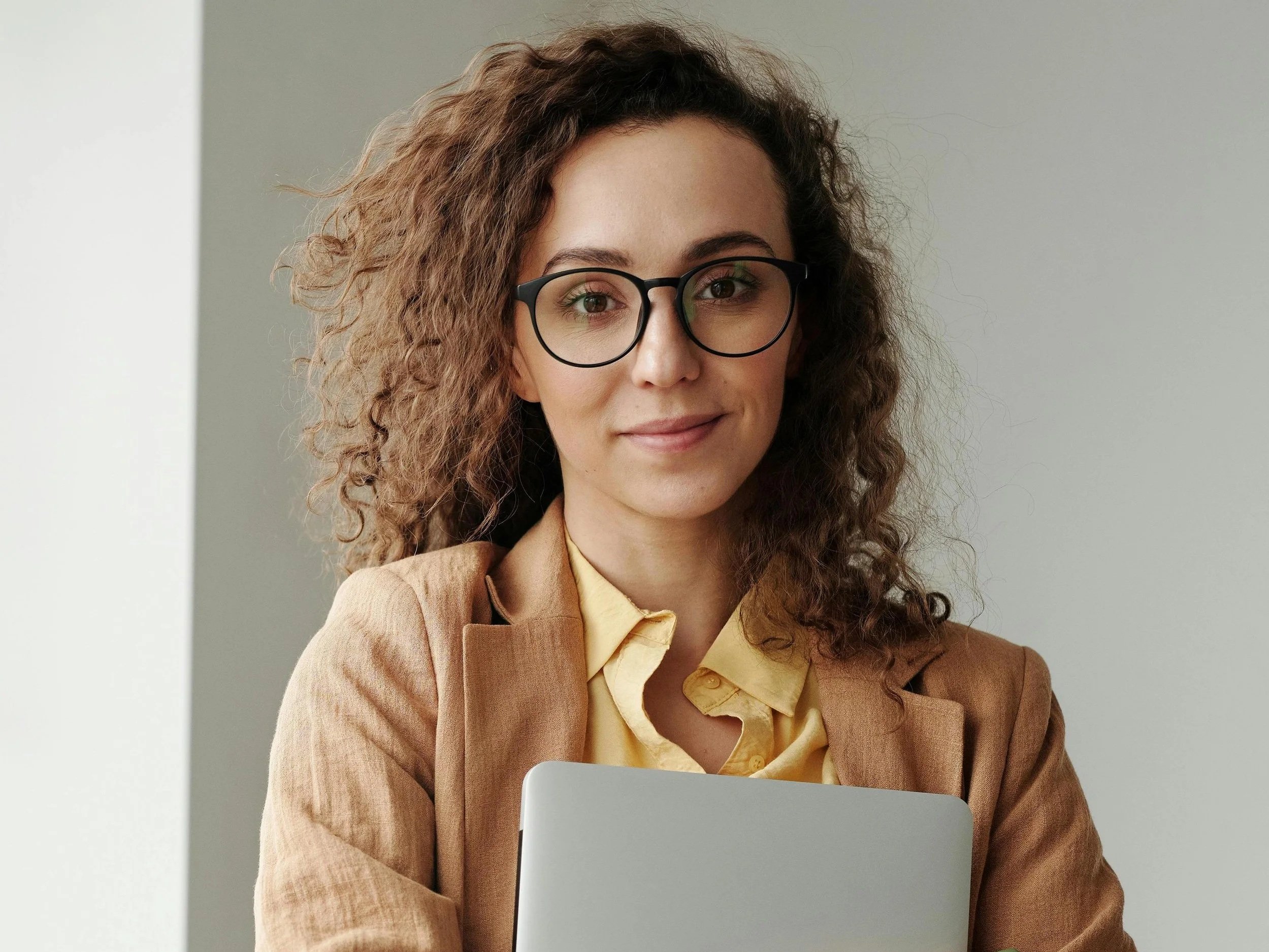 A woman in business attire sitting in front of a laptop, smiling, with curly hair and glasses.