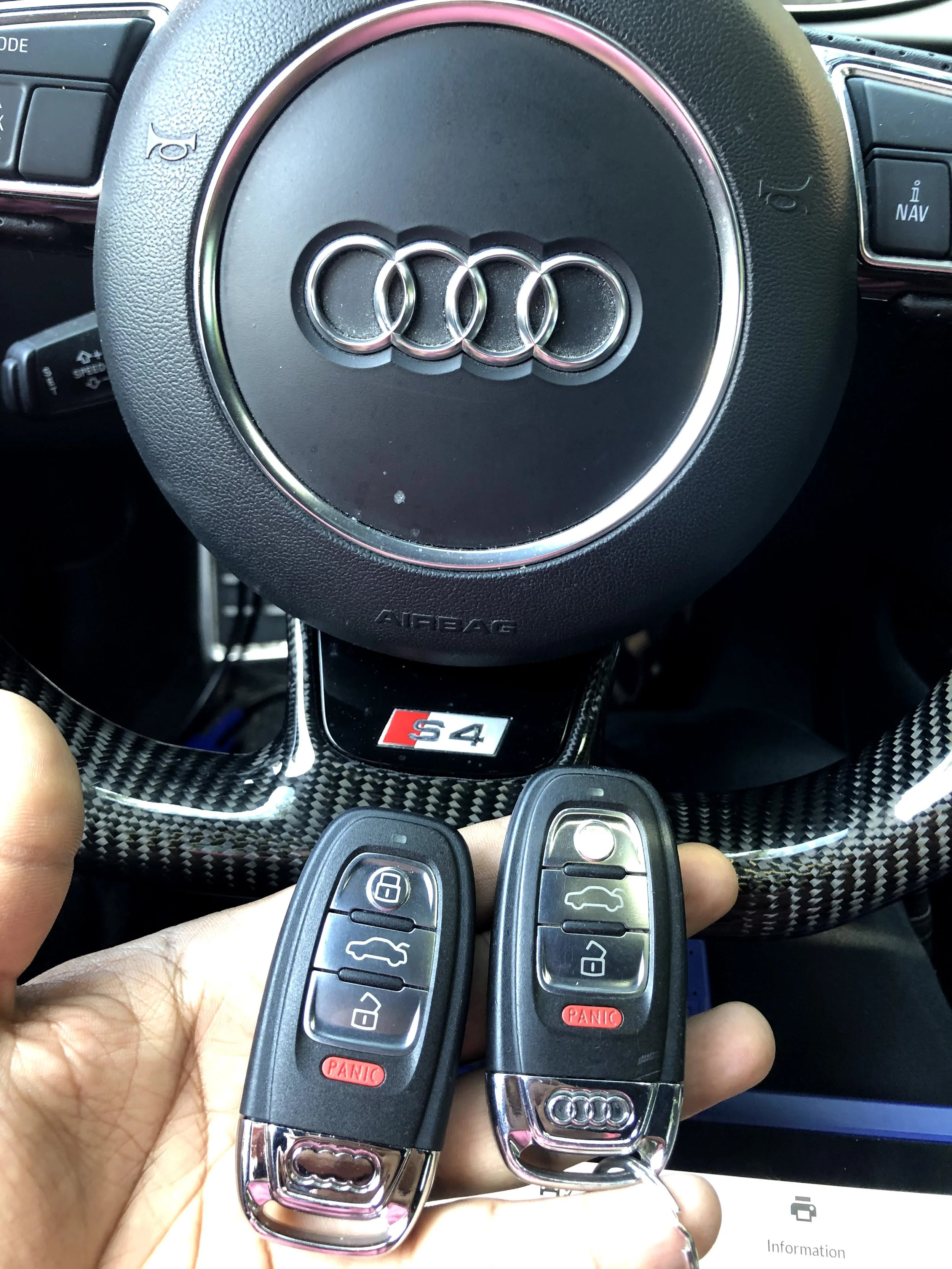 A close-up photo of an Audi steering wheel with the Audi logo in the center. In the foreground, a hand is holding two Audi key fobs with black and silver design and red panic buttons.