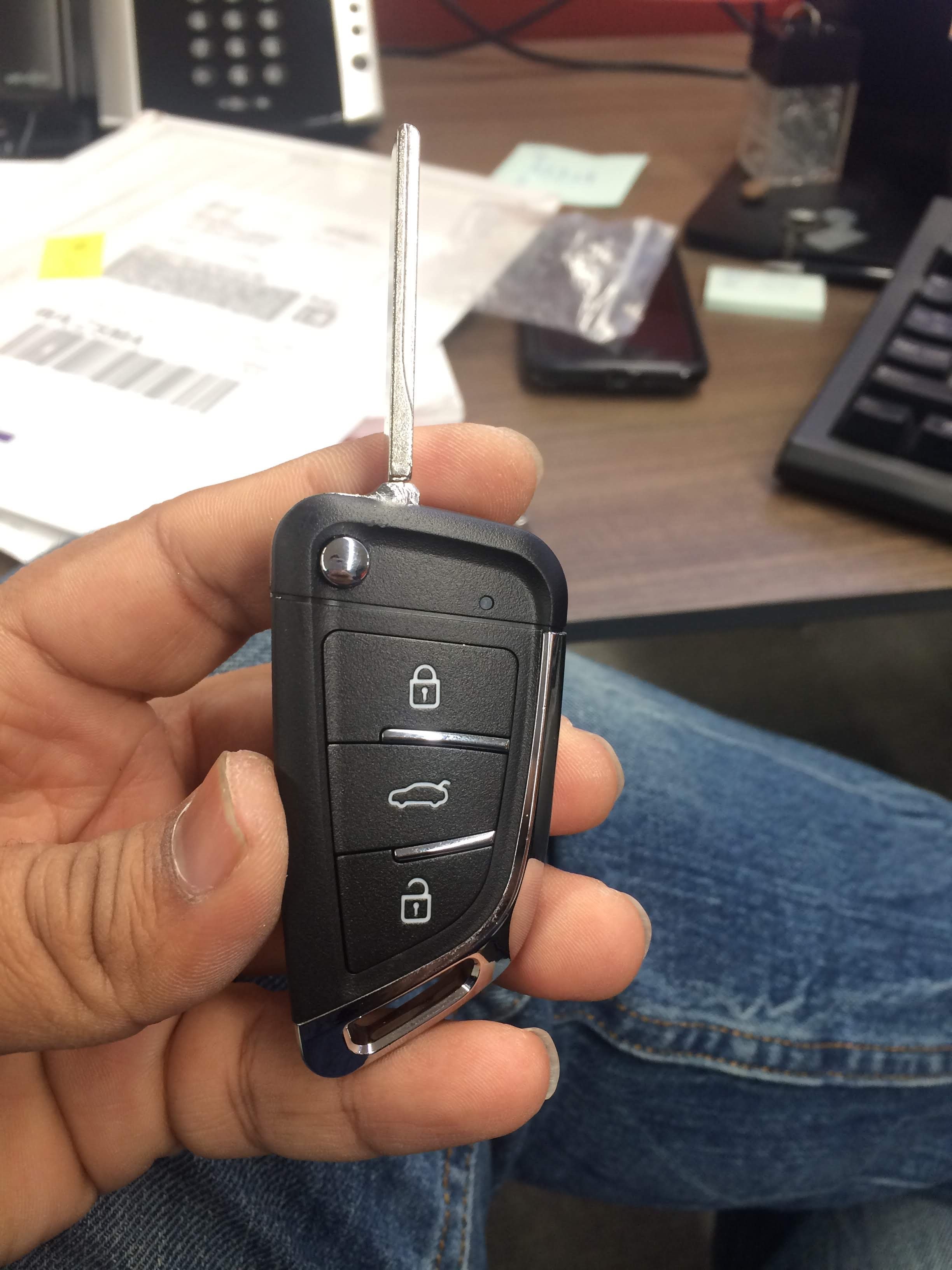 A person holding a car key with a black plastic key fob and metal key blade. The background shows a cluttered desk with papers, a smartphone, a keyboard, and various office supplies.