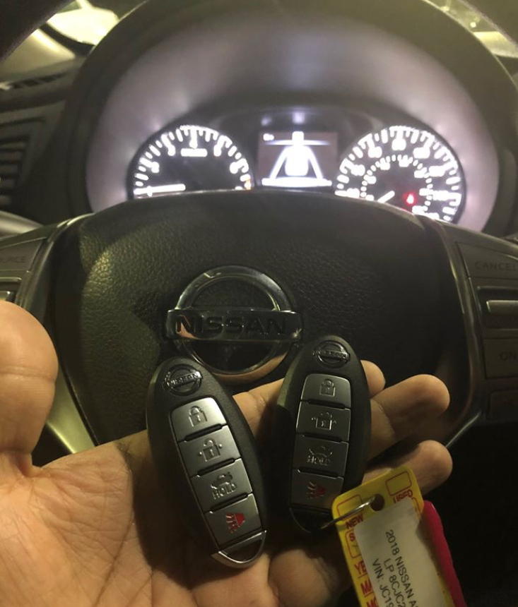A person holding two Nissan car key fobs inside a car, with the Nissan logo visible on the steering wheel and the dashboard showing the speedometer and other gauges.