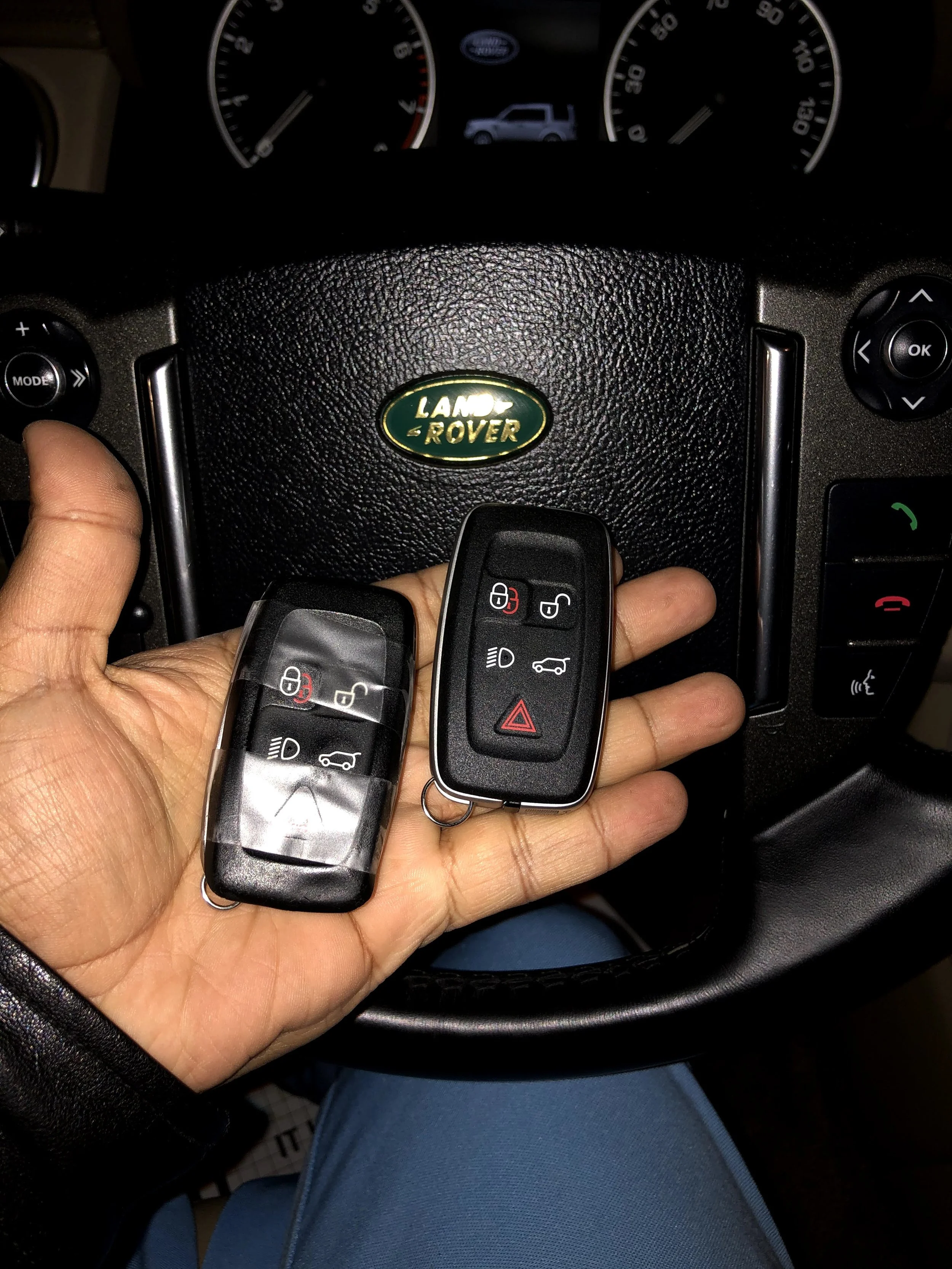 A person holding two car keys over a car steering wheel with a Land Rover badge, with the car dashboard visible in the background.