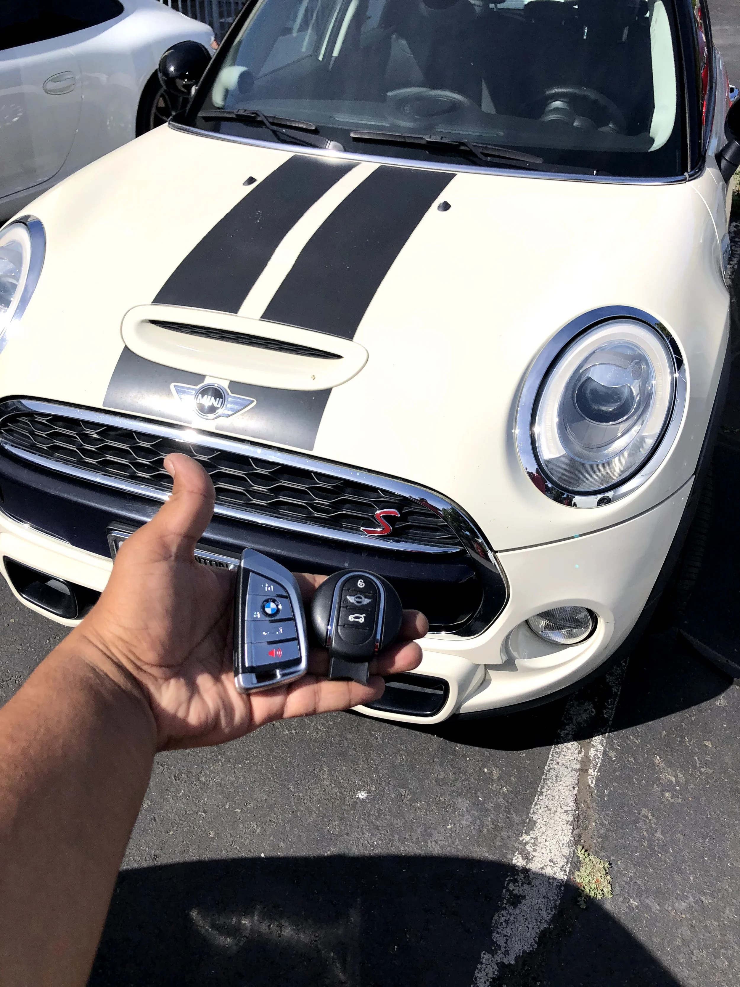 A person holding a BMW key fob with a Mini Cooper car in the background. The car has a white exterior with two black racing stripes on the hood.