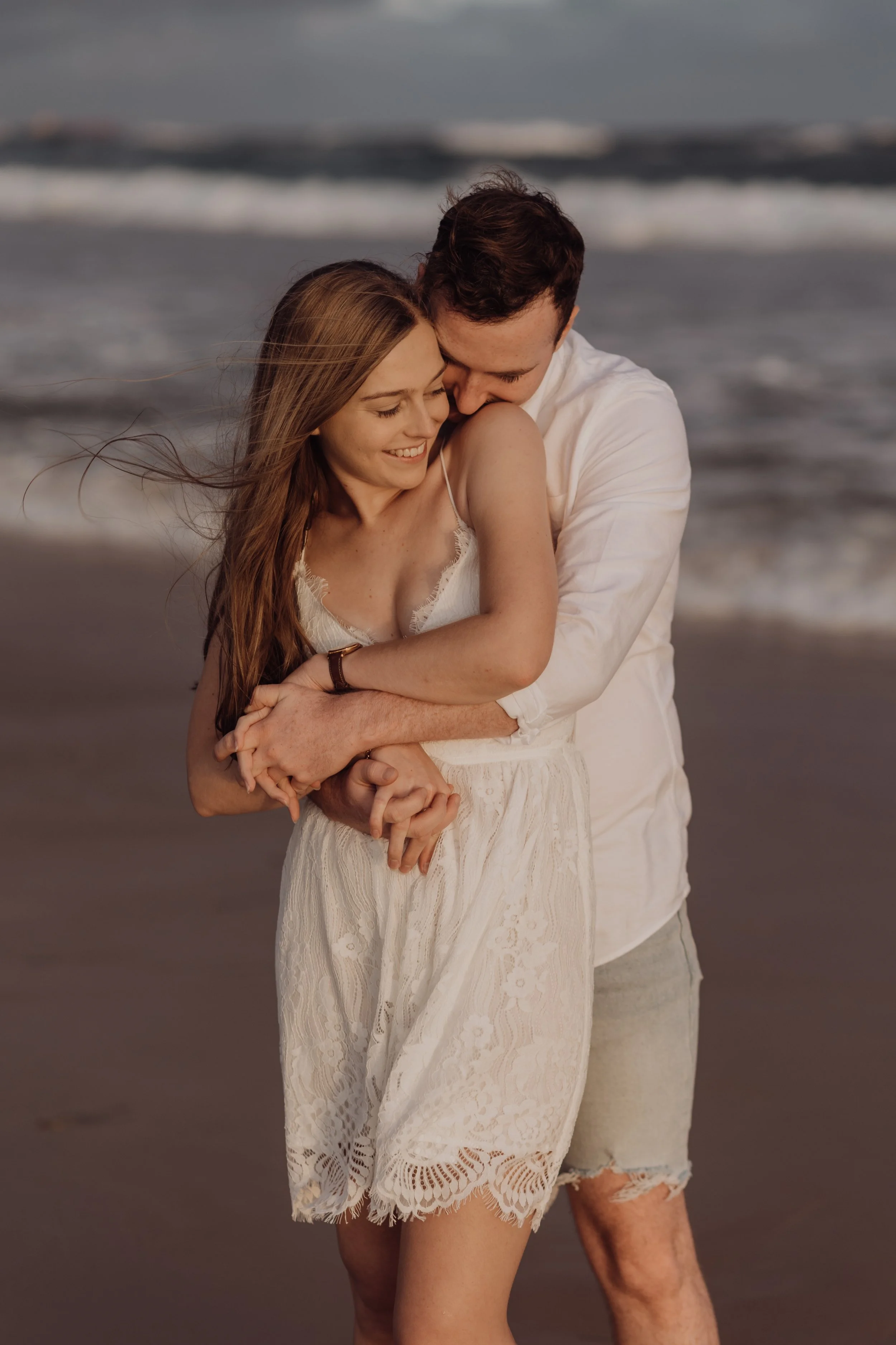 A happy couple embracing on the beach during sunset, with the ocean and waves in the background.