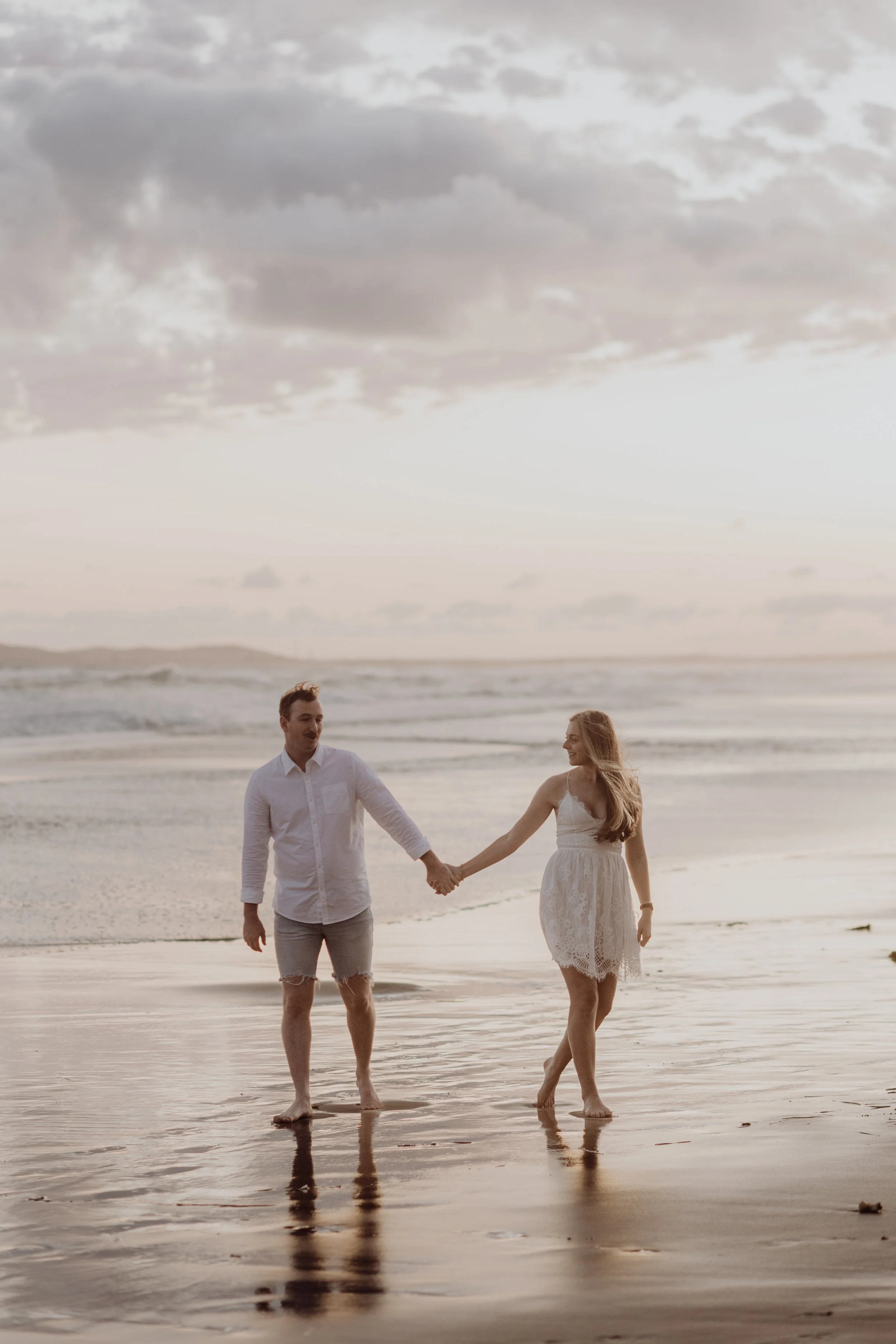 A couple holding hands walking together on a beach during sunset, with the ocean in the background and cloudy sky above.