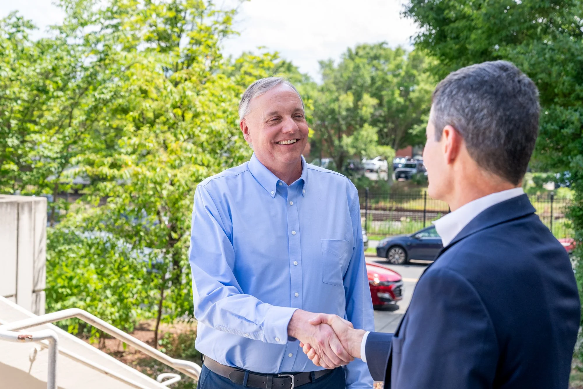 Two men shaking hands outdoors with trees and parked cars in the background.
