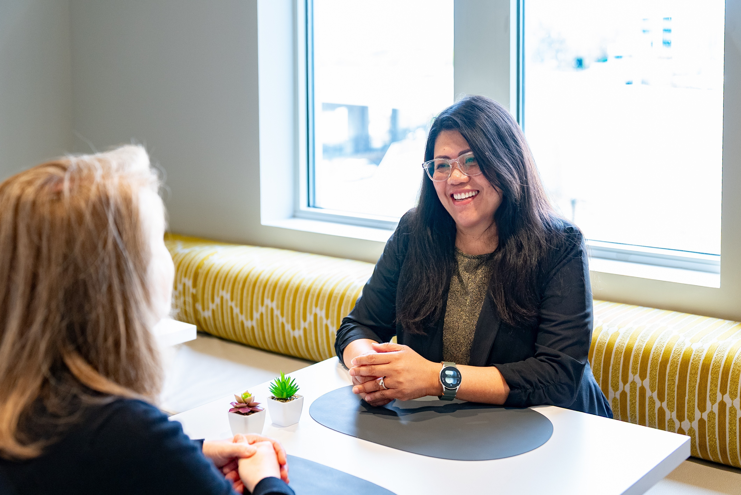 Two professionals smiling and talking across a small table in a bright office setting.