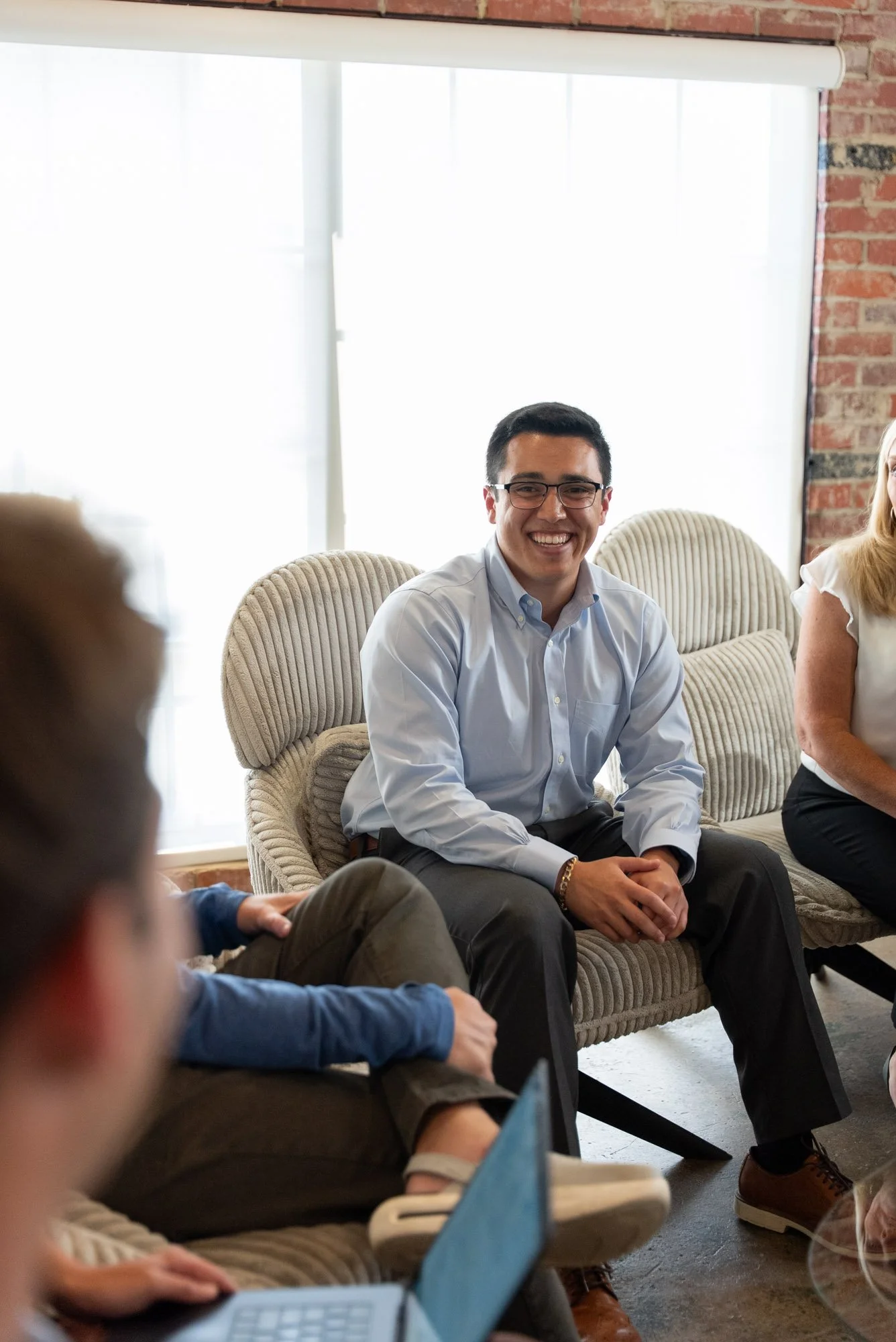 A group of people sitting on a beige couch in a room with large windows and brick walls, engaged in conversation. One man in a blue shirt and glasses is smiling.