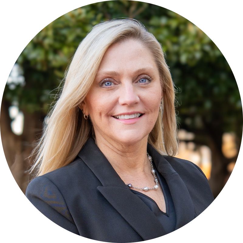 A smiling blonde woman with blue eyes wearing a light blue top, earrings, and a necklace, outdoors with blurred greenery in the background.