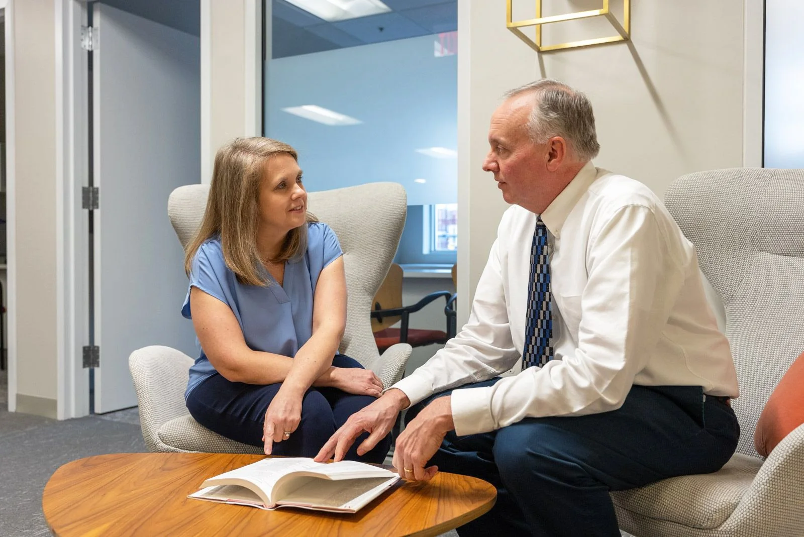 A woman and a man seated, pointing at a book in an office setting.