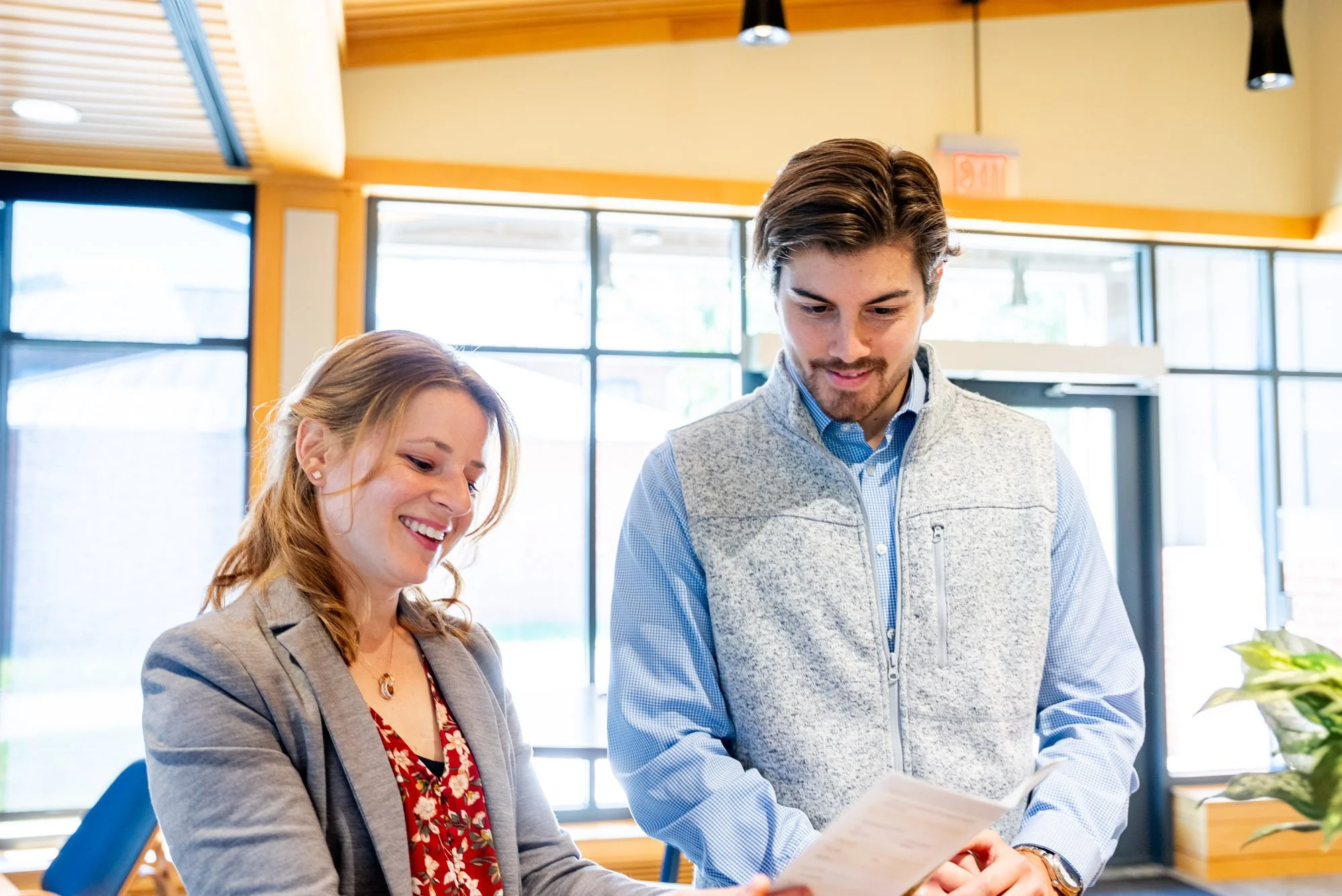 A man and woman standing inside a bright, modern office or cafe, smiling and looking at a document together.