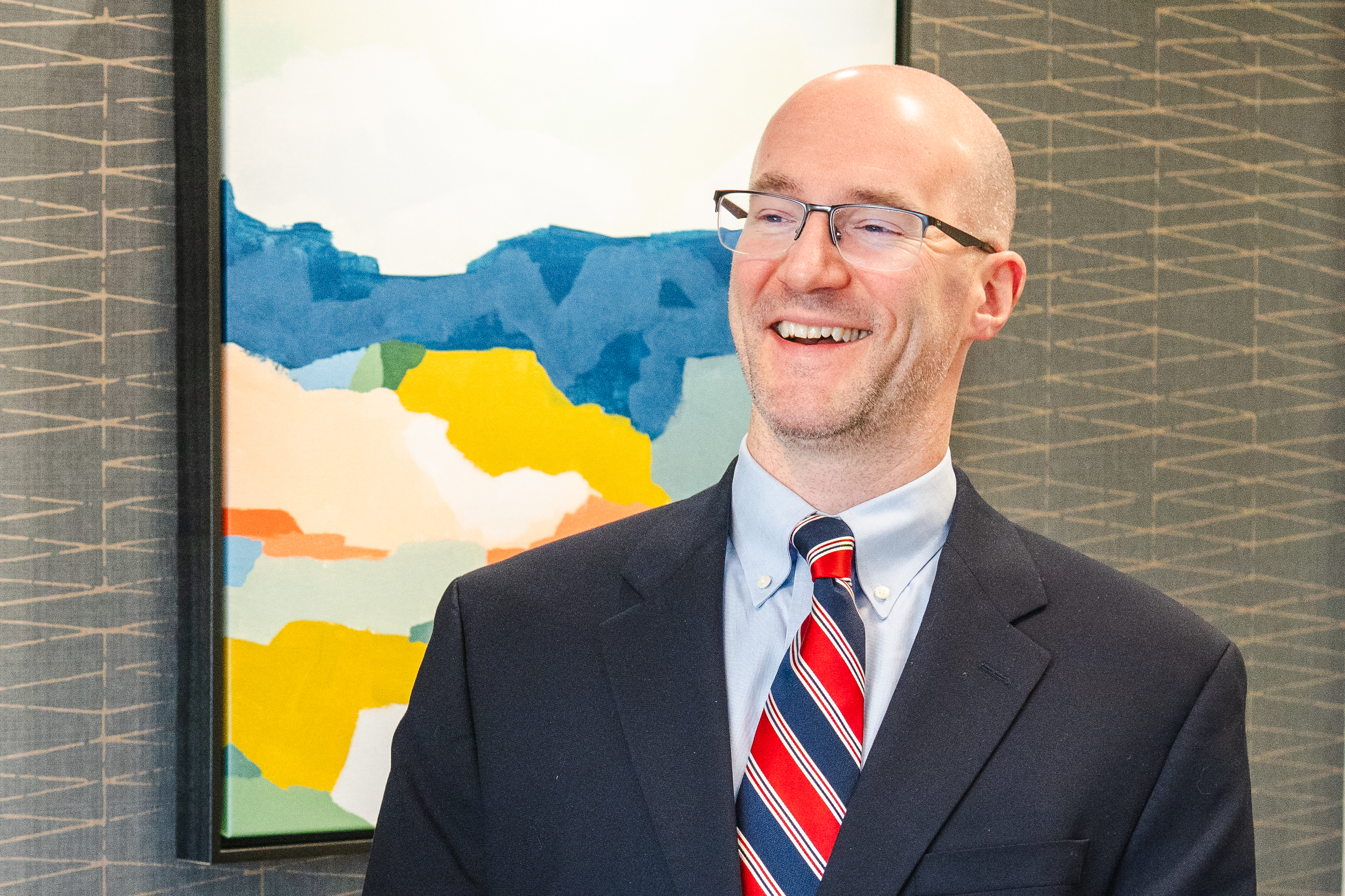 Smiling tax professional wearing glasses, a navy suit, and a red striped tie, standing indoors in front of a colorful abstract wall art display.