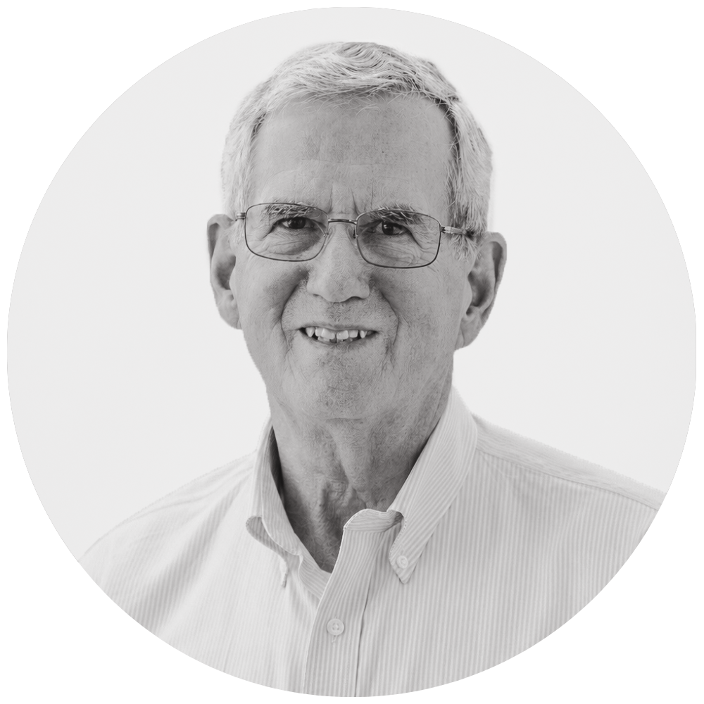 Portrait of an older man with gray hair and glasses, wearing a white button-up shirt with the logo 'Hantzm Wieb' on it, outdoors with trees and sunlight in the background.