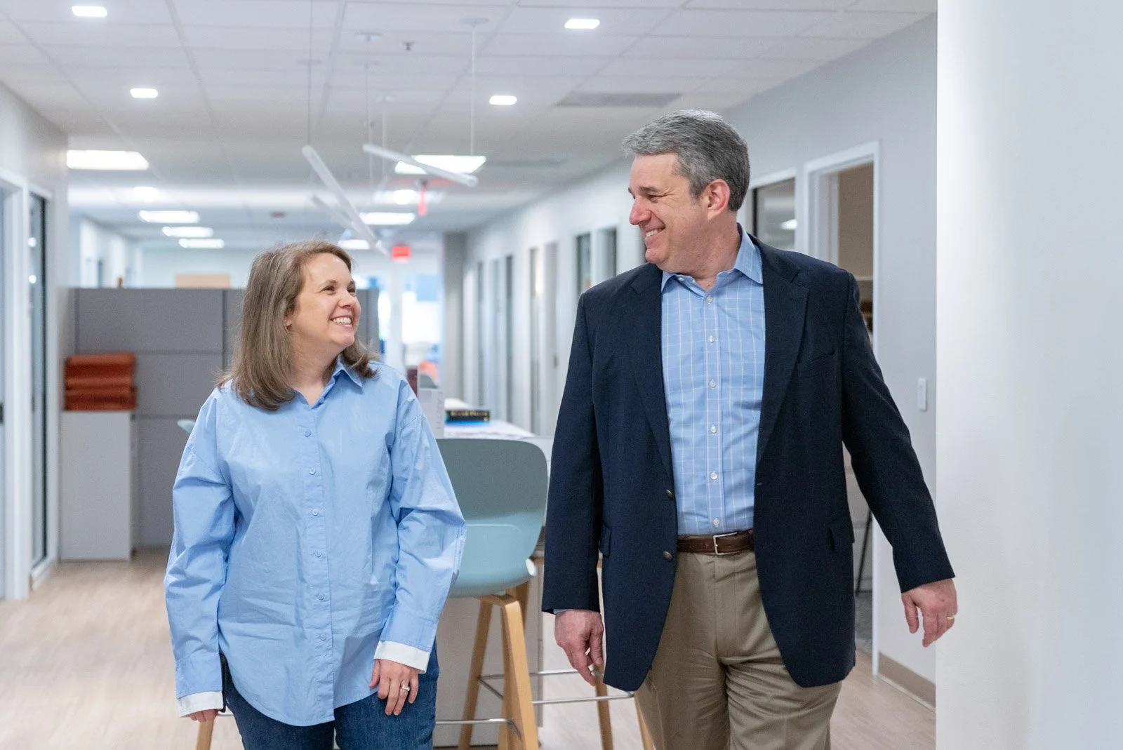 A man and woman walking down a hallway, smiling at each other in an office environment.