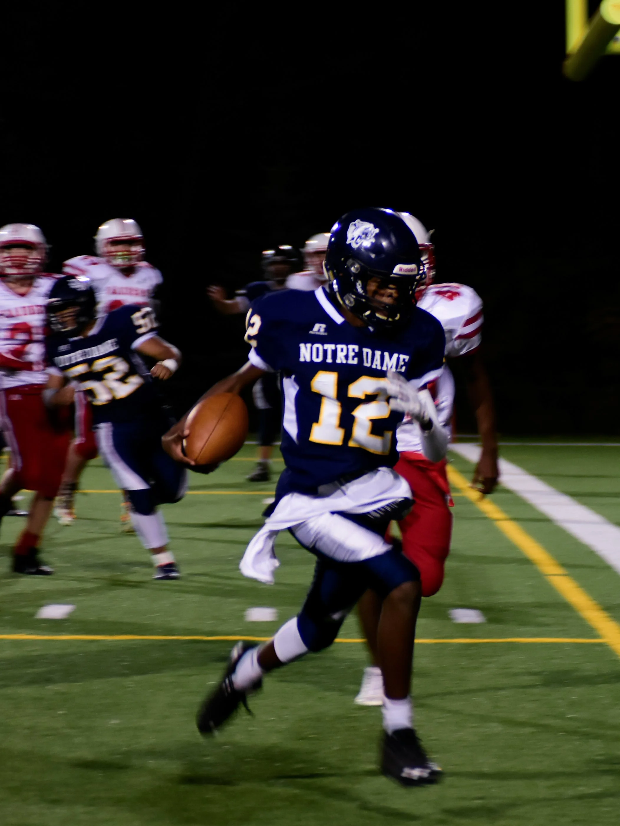 A football player from Notre Dame wearing number 12 running with the football during a game under the night sky, surrounded by opponents in red and white uniforms.