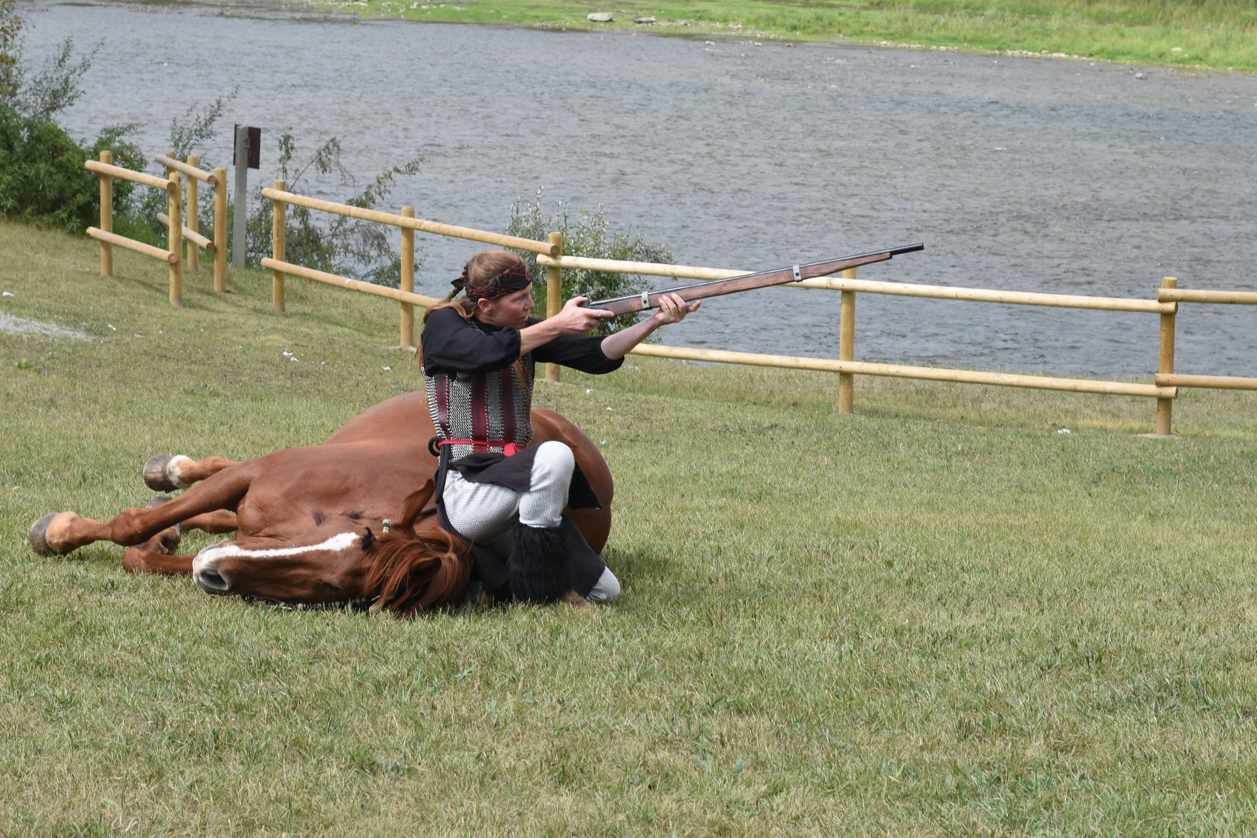 A person in historical clothing riding a rearing brown horse while aiming a long rifle near a body of water and a wooden fence.