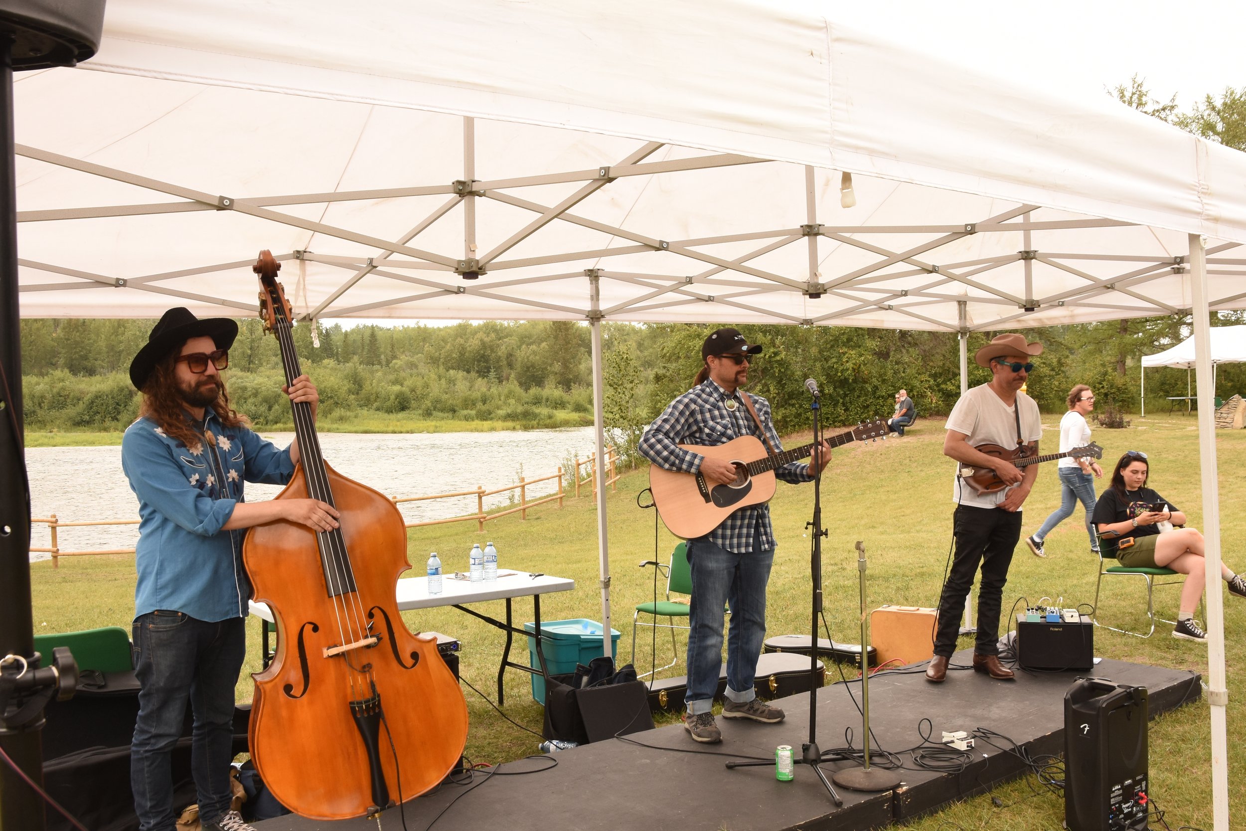 A band of four musicians performing outdoor under a large white tent near a lake. One plays a double bass, another plays an acoustic guitar and sings into a microphone, the third holds a mandolin, and the fourth is seated, looking at her phone.