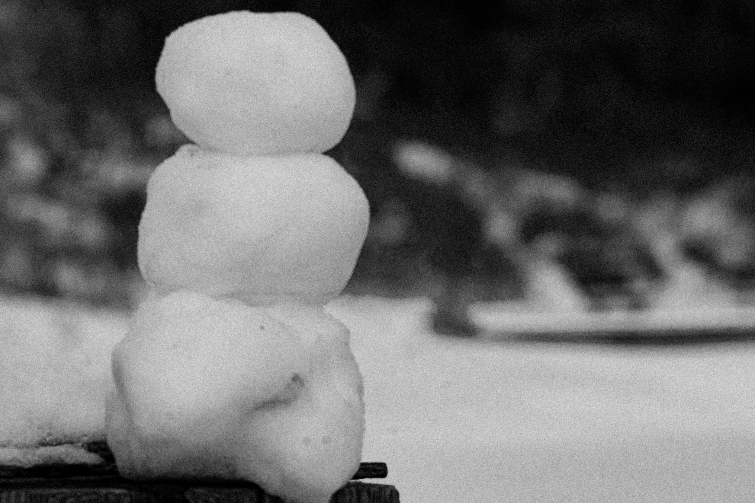 Close-up of a small snowman made of three snowballs stacked on top of each other, on a wooden surface, with a blurred background.